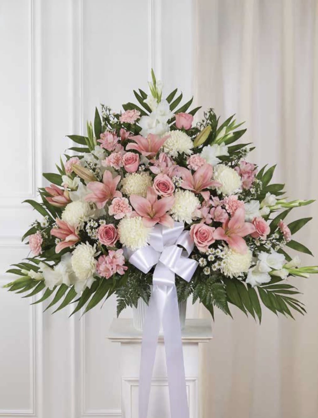 White and Pink Pedestal Arrangement - A graceful pedestal arrangement of pink and white blooms makes an immediate, uplifting impression-perfect as a sympathy piece or an elegant celebration bouquet. This white and pink pedestal arrangement pairs pink lilies and roses with white chrysanthemums, carnations, and delicate alstroemeria to create a soft, layered presentation that reads both ceremonial and celebratory. The balanced, symmetrical shape and white ribbon bow give the design a polished, respectful presence for memorial services or a meaningful delivery to someone you want to honor.  The arrangement sits in a white vase set into a large, formal pedestal-style design, with abundant green foliage to frame the blooms and add fresh contrast. Pink lilies provide height and fragrance, while roses and alstroemeria add texture and romantic color; chrysanthemums and carnations fill the composition for fullness and longevity. Its broad spread makes it visible from a distance and suitable for placement beside a casket, at a reception space, or as a standout piece in a home.  Choose this arrangement when you want the recipient to feel genuinely seen-whether offering condolences, celebrating a milestone, or sending a thoughtful sympathy or occasion gift. Because of its size and formal construction, please note it's intended for delivery and display rather than mail; contact us for delivery timing and placement guidance to ensure it arrives looking its best.