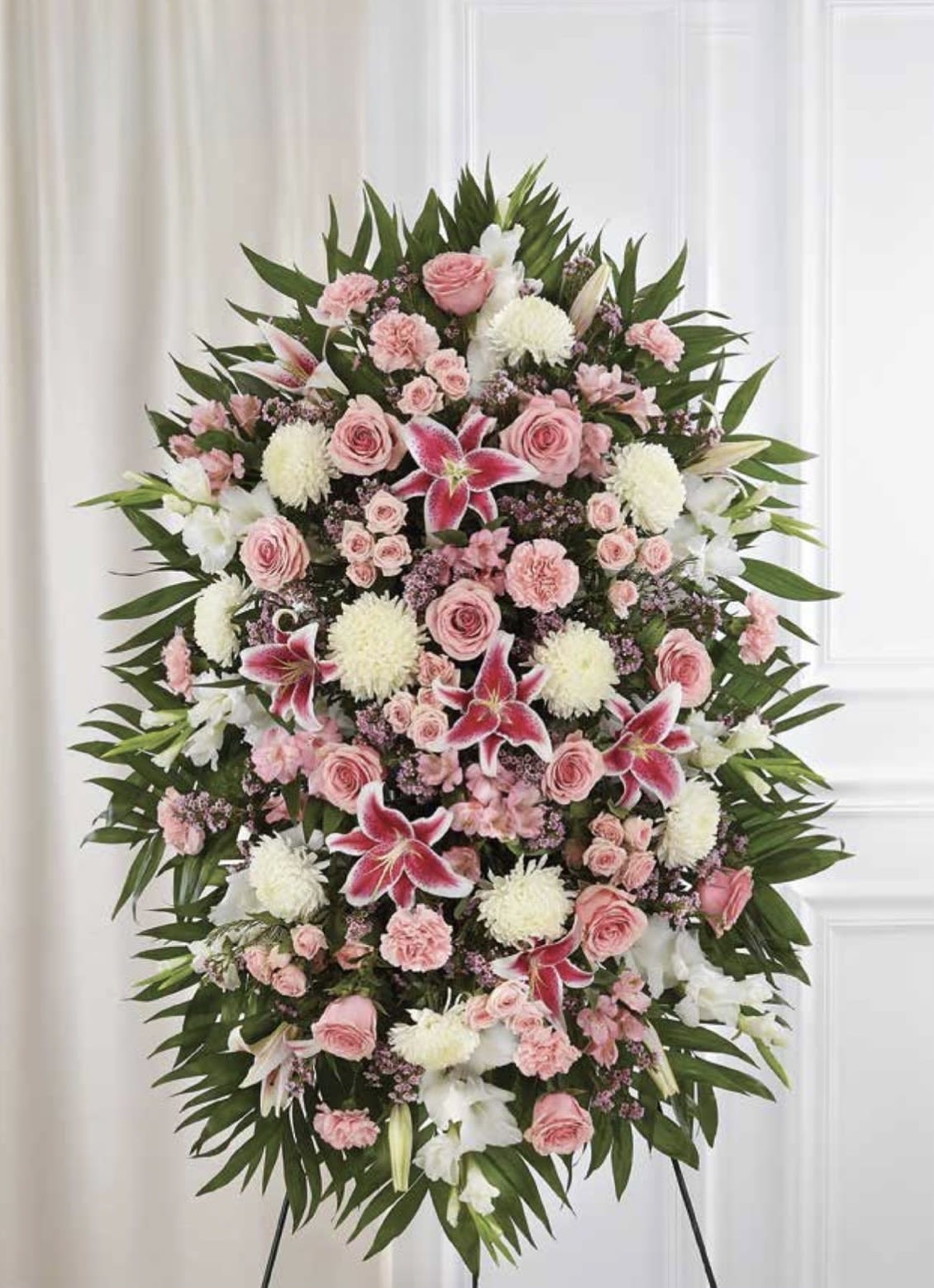 White &amp; Pink with Stargazer Lilies Standing Spray - A graceful sympathy standing spray that reads as both serene and uplifting, perfect for a memorial service or to express condolences. This flower arrangement for funerals features a formal oval silhouette on a standing easel, combining white and pink blooms to convey gentle comfort while the dramatic stargazer lilies provide a striking focal point.  The spray is densely layered with pink roses and carnations nestled among white chrysanthemums, alstroemeria, and lilies, all backed by deep green foliage that forms a strong outer frame. Mounted on a floral spray frame and standing easel, the piece presents a polished, lasting tribute with rich texture from the mixed petals and the sculpted oval shape.  Choose this arrangement to honor a loved one or to send sympathy to the family-its colors and composition express compassion and respect without being showy. Sized as a large standing spray, it arrives ready to display at a service; keep flowers in a cool place and ensure the oasis or foam stays hydrated while on site for best longevity.