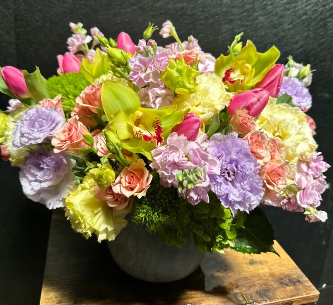 Golden Bloom - A lively, garden-style flower arrangement that reads joyful and celebratory, perfect as a birthday bouquet, spring centerpiece, or thoughtful thank-you gift. Golden Bloom pairs bright pink tulips with lime-green orchids and soft pastel roses and carnations so the person you're sending it to feels truly seen-cheerful, uplifted, and celebrated in equal measure.  This dense, low rounded arrangement is packed with lavender, blush, cream and pale yellow blooms layered tightly for an abundant, textured look. Ruffled carnations, smooth tulip buds, orchid faces and mixed seasonal flowers create a playful yet elegant mix of forms and colors. It arrives arranged in a matte gray ceramic vase that complements the vivid chartreuse accents and makes the bouquet ready to display.  Ideal for spring gifting, birthdays, anniversaries, or as a bright pick-me-up, Golden Bloom looks lovely on a dining table, entry console, or office desk. No special care is required beyond fresh water-place in a cool spot away from direct heat and enjoy the layered colors for days as stems open.