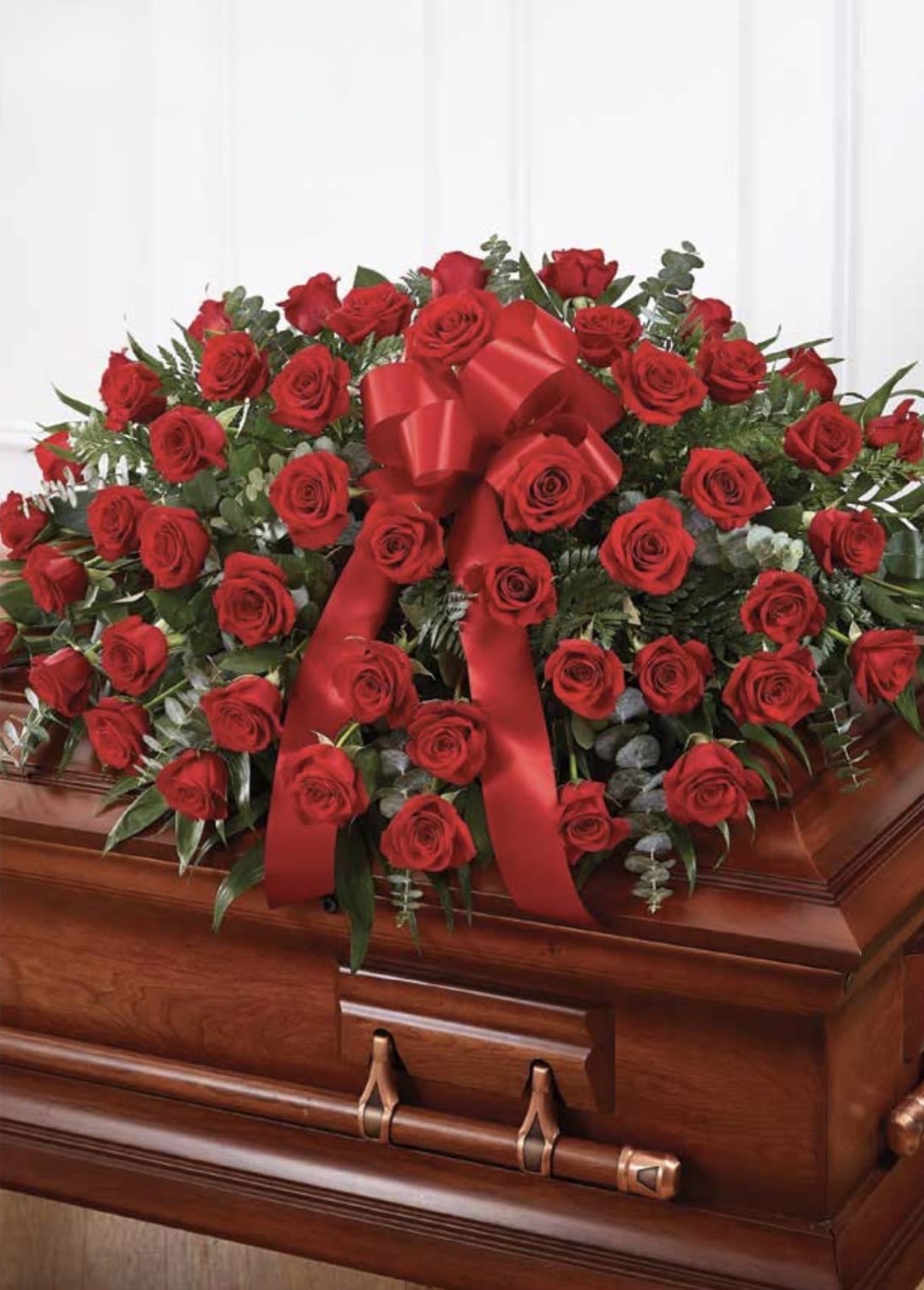 Red Rose Casket Spray - A lush cascade of deep red roses laid across a polished wooden casket creates a solemn, reverent statement - a red rose casket spray designed for funeral and memorial services. The classic red roses and greenery convey love and respect, making this arrangement a meaningful way to honor the person you're thinking of and to express sympathy at a time of loss.  The spray is dense and formal: long-stemmed red roses are arranged in a broad, low composition that rests across the casket, accented with mixed greenery for texture and depth. A prominent satin red ribbon is tied through the center, adding a ceremonial touch and completing the traditional, elegant presentation. Sized and shaped to drape gracefully over a standard casket, it presents a unified red-and-green palette with touches of white from the greenery highlights for a dignified finish.  Appropriate for a funeral, memorial, or graveside service, this arrangement speaks of enduring love and respect for the deceased. Please let us know service details and delivery timing when ordering so we can ensure careful placement on the casket; our team handles delicate arrangements with the care and discretion these moments require.