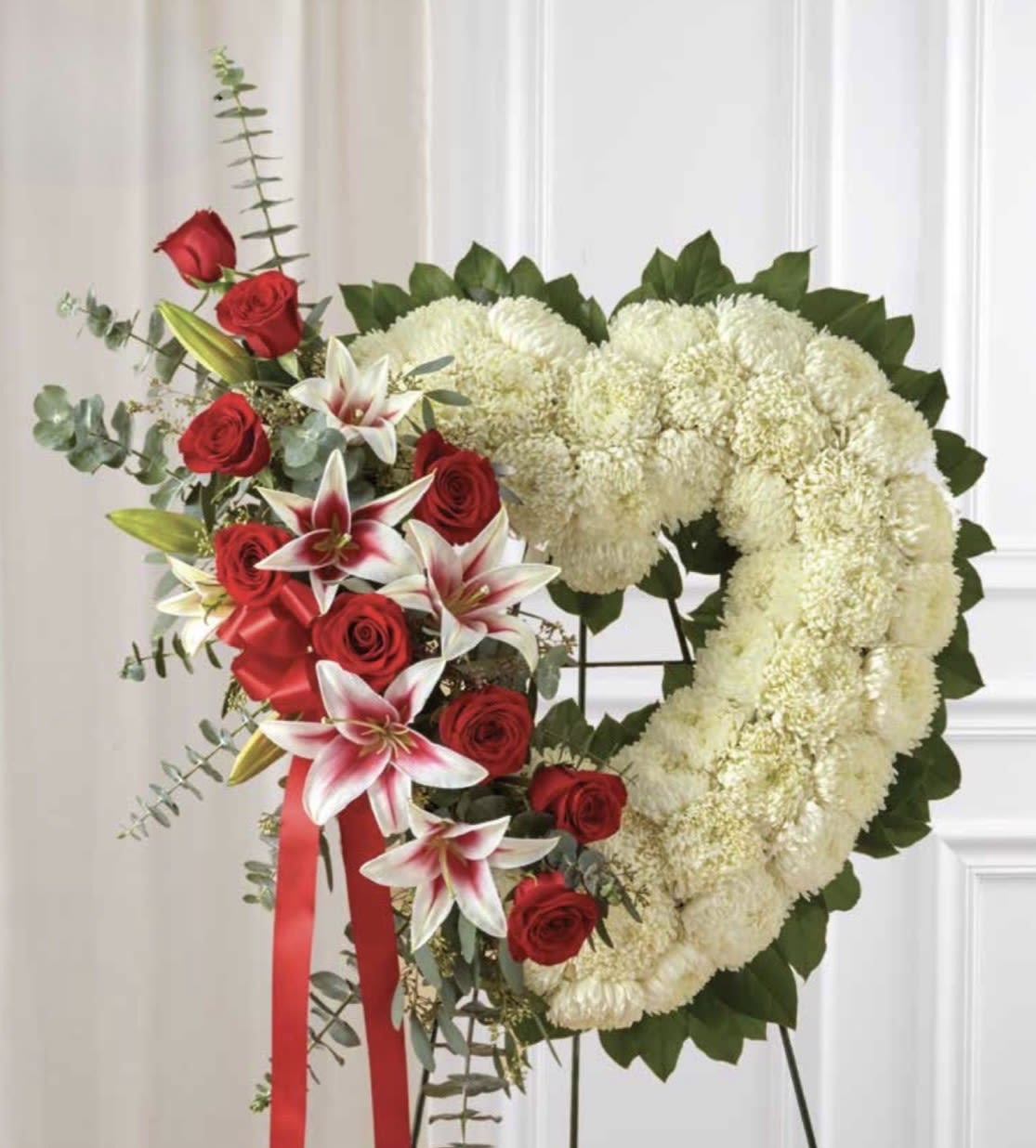 White Heart with Rose &amp; Stargazer Cluster - A large heart-shaped sympathy arrangement centered on pure white chrysanthemums or carnations makes a powerful, consoling statement for a memorial or funeral service. The white heart with rose &amp; stargazer cluster offers a formal, respectful tribute the person you're thinking of deserves, while the red roses and lilies introduce a warm, personal note that speaks of love and remembrance.  Dense white chrysanthemums or carnations form the heart and are neatly outlined with deep green foliage for clear definition and visual contrast. A clustered spray of red roses and stargazer lilies with soft pink centers sweeps down the left side, adding color and texture against the white. The arrangement is mounted on a standing easel and finished with a long red ribbon for a ceremonial presentation that reads both classic and heartfelt.  Appropriate for a funeral service, memorial or as a graveside tribute, this piece is designed to comfort and honor-she or he will feel cherished and remembered. The easel-mounted format makes placement at a service simple; no additional care is required beyond standard handling at a ceremony.