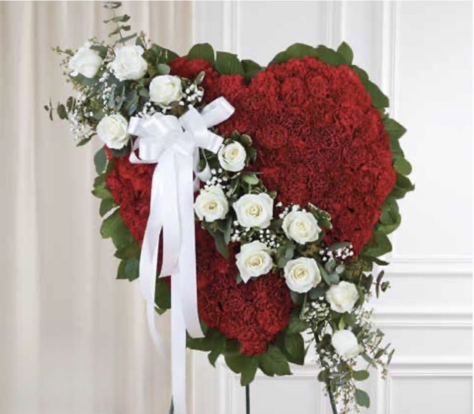 Solid Red Heart with White Rose Cluster - A bold heart of deep red carnations arranged on a standing easel makes an immediate, heartfelt statement-perfect as a sympathy tribute or memorial heart that honors a life with dignity. The diagonal cluster of pristine white roses and small white filler flowers draws the eye across the design, while a white ribbon bow near the top-left adds a formal, respectful touch that signals condolence and remembrance.  This floral arrangement combines dense red carnations for rich color and texture with a contrasting spray of white roses to provide visual relief and symbolic purity. Greens peek through the composition to frame the heart and add depth; the piece is securely mounted on a standing easel for display at a service or graveside. The overall scale is substantial and designed to read clearly from a distance, offering a full, dramatic silhouette.  Made to express sympathy and honor, this standing heart is the kind of tribute that helps the person you're remembering feel seen and celebrated. Please indicate service date and delivery details at checkout so we can ensure timely placement; standard care is minimal while on the easel-keep out of direct sun and mist lightly if needed to maintain freshness before the program.