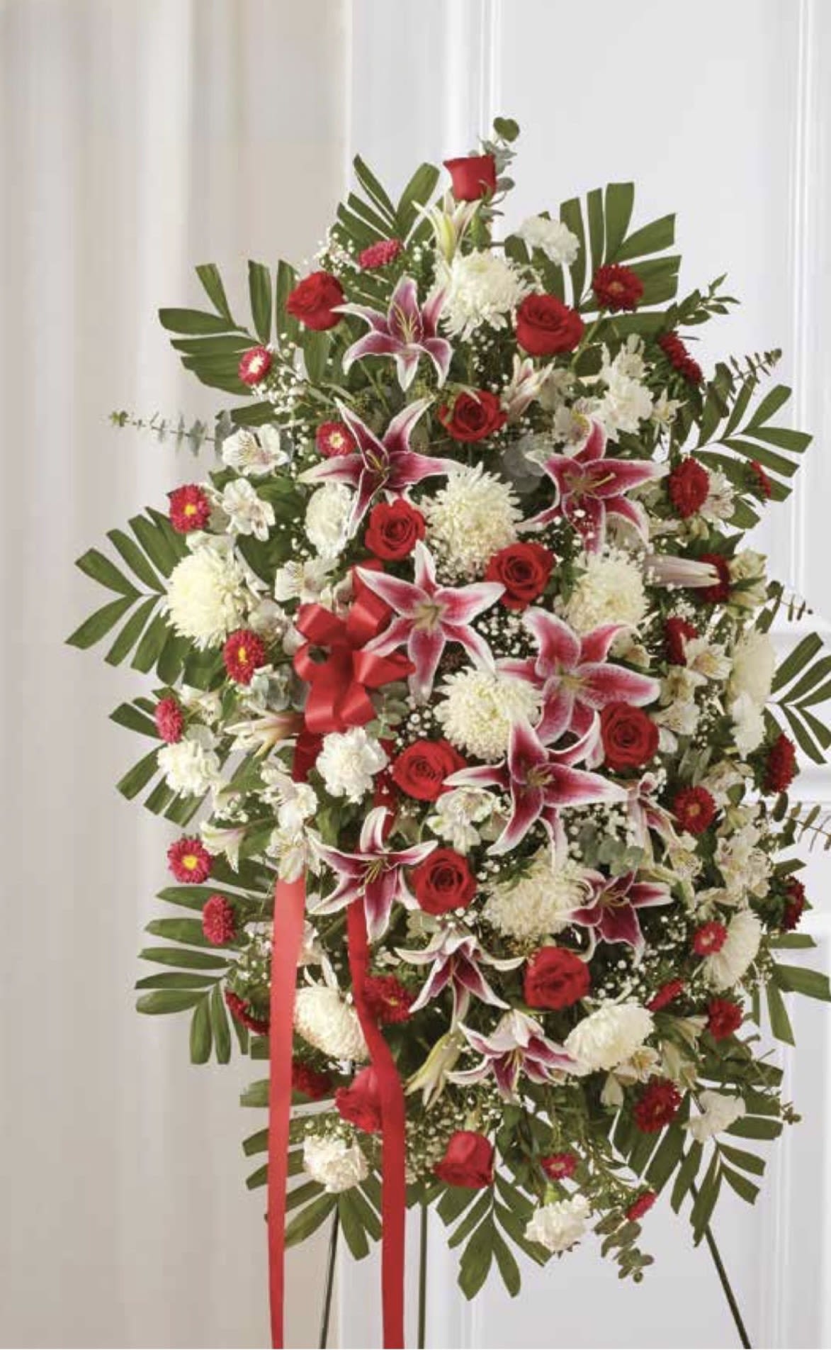 Red &amp; White with Stargazers Lilies Standing Spray - A dignified standing spray of red and white designed to honor a loved one - perfect for a sympathy or memorial service. The arrangement pairs deep red roses with crisp white blooms and striking pink-and-white stargazer lilies, creating a classic red &amp; white standing spray that reads as both respectful and celebratory for the person you're remembering.  This large spray is arranged in a rounded vertical form with dense, layered clusters of roses, lilies, carnations and chrysanthemums. Broad tropical-style greenery frames the edges for a full, formal silhouette while red ribbon streamers and an easel hard-good give it a ceremonial finish. The mix of red, white, pink, and green delivers contrast and softness: the stargazer lilies add texture and fragrance amid the velvety roses and cushiony carnations.  Choose this arrangement when you want the recipient's family to feel comforted and seen - it's the kind of tribute that conveys deep respect and heartfelt sympathy. Sized as a full standing spray on an easel, it arrives ready to display; stems are arranged for longevity, and local delivery ensures it reaches the service promptly.