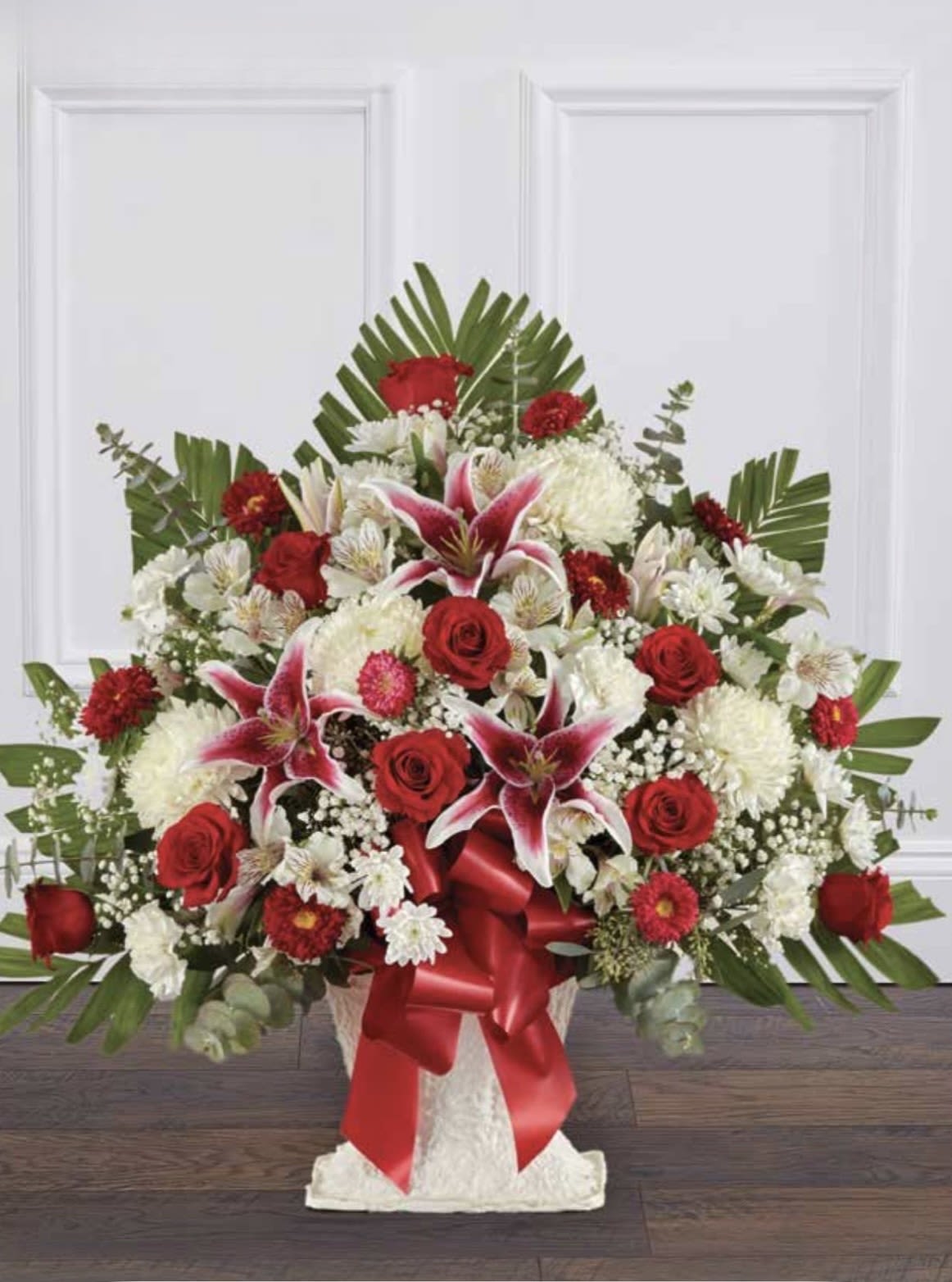 Red &amp; White with Stargazer Lilies Pedestal Arrangement - A bold, fan-shaped pedestal arrangement centered on red roses and pink stargazer lilies, perfect as a romantic centerpiece, anniversary tribute, or elegant sympathy display. The red and white color story reads strong from a distance and intimate up close-the lilies' fragrant blooms and the deep, velvety roses create a dramatic first impression that will make the recipient feel deeply noticed and honored.  This large, symmetrical design layers white chrysanthemums and small daisy-like blooms among lush green palm fronds and eucalyptus-like foliage for texture and movement. Arranged in a classic white vase and finished with a wide red satin ribbon, the composition measures as a full, sculpted silhouette meant to stand on a pedestal or entry table. The mix of blooms provides contrast in shape and scent-starry lilies, rounded roses, and soft mums-so the arrangement looks beautiful from every angle.  Ideal for marking milestones, offering condolences, or celebrating love, this arrangement sends a clear message of respect and affection. Order with standard care: keep water fresh, trim stems if needed, and place away from direct heat to extend life; its substantial size and formal ribboned vase make it a showpiece arrival for the person you want to honor.