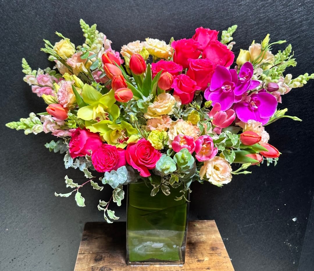 Mia's Signature Garden - Lush waves of hot pink roses and vibrant purple orchids make an immediate impression in this signature flower arrangement-perfect as a birthday bouquet, congratulations gift, or romantic surprise. The composition reads celebratory and full of life: the person you're thinking of will feel seen and cherished when greeted by these bold, joyful colors.  Closely layered across the top of a tall green glass vase, coral tulips, yellow-green orchids, soft peach blooms, and pink carnations create a dense, garden-like crown, while airy snapdragon spikes and variegated foliage add height and movement. The mix of intense hot pinks and purples with gentler peach and yellow tones gives strong visual contrast and a lively, textured look that suits both cheerful milestones and romantic moments.  Designed to feel abundant on delivery, the arrangement arrives ready to display in its green glass vase; keep flowers in fresh water away from direct heat and change the water every two days to extend bloom life. Choose this for someone who delights in bold color and lush, sculptural floral moments-the kind of bouquet that makes them feel truly celebrated.