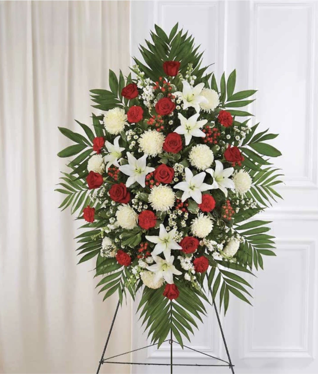Red and White Standing Spray - A formal red-and-white standing spray that commands attention with its dramatic palm-frond frame-perfect as a sympathy arrangement or memorial tribute. The bold contrast of red roses and pure white lilies creates a dignified focal point, while round white chrysanthemums and delicate filler blooms soften the composition, giving the person you're honoring a sense of reverence and warmth.  This large standing spray is arranged in a dense, symmetrical silhouette on a metal easel for prominent display at a funeral service or graveside memorial. Deep green palms and foliage provide a sculpted backdrop to clusters of red roses, open white lilies, and cushion chrysanthemums, producing a polished, traditional look that reads clearly from a distance.  Choose this spray when you want the recipient's memory to be honored with solemn beauty-the kind of tribute that feels respectful and meaningful to family and friends. The piece arrives ready to present on the included standing easel; as with all fresh floral sprays, keep it sheltered from extreme heat and wind for best longevity.