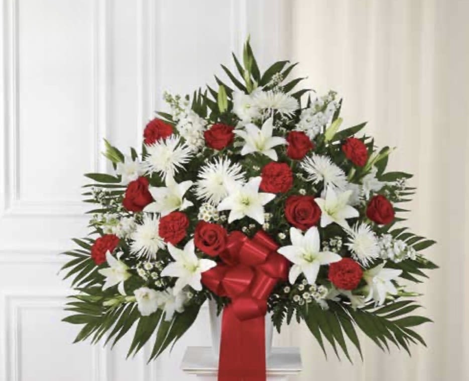 Red and White  Pedestal Arrangement - A large, formal standing flower arrangement that commands attention-perfect as a sympathy pedestal or for a solemn holiday display. This red and white pedestal arrangement pairs classic red roses and carnations with pure white lilies and spider chrysanthemums to create a polished, symmetrical statement piece that reads both respectful and celebratory for memorial services, funeral home viewings, or ceremonial occasions.  Dense layers of blooms form a broad, upright silhouette set against dark green foliage for strong contrast and visual depth. The design is finished with a red satin ribbon and presented on a clear vase base so the arrangement reads as a display rather than a handheld bouquet; the scale and structure make it ideal for placement at an altar, beside a casket, or as a focal tribute. Recipients will feel honored and remembered by the thoughtful, traditional composition-order with any special message for delivery details and placement instructions.