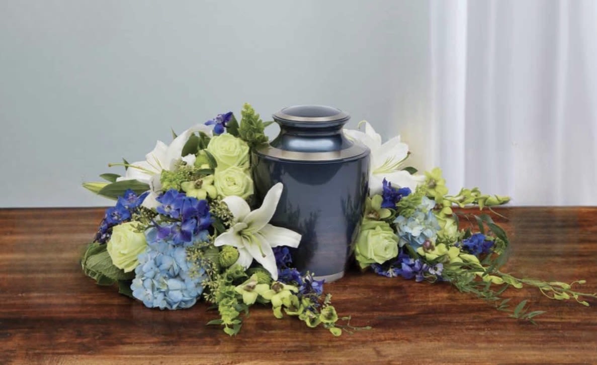 Blue and White Crescent Cremation Piece - A cool, serene crescent of blooms cradles the urn to create a respectful memorial piece perfect for a cremation service or sympathy tribute. The low, spreading design keeps the focus on remembrance-this cremation arrangement reads as calm and dignified, the kind of floral expression that helps the person you're honoring feel quietly celebrated.  White lilies, pale green roses, and blue hydrangeas form the heart of the composition, joined by clusters of soft blue blooms for gentle contrast and depth. The flowers are arranged symmetrically in a low ring that extends outward across the table rather than rising tall, creating a full, balanced profile in white, green, and blue tones.  Appropriate for a funeral home, memorial service, or as a centerpiece at a remembrance gathering, this piece offers a composed, consoling presence for family and friends. Designed to be low and wide, it's intended to sit on a service table or memory table; minimal care is needed once set in place-keep cool and out of direct sunlight to maintain freshness during the service.
