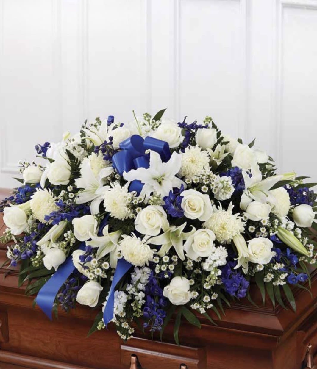 White with Blue Casket Spray - Pure white roses and lilies form the calm, dignified heart of this funeral casket spray, accented with deep blue and purple blooms to add depth and reverence. As a casket spray for memorial services, it offers a formal, traditional expression of sympathy-perfect for conveying respect and honoring the person you're thinking of.  This large, low arrangement sits across the casket with a dense, layered surface of fuller round chrysanthemums, daisies and small filler flowers that create rich texture and visual weight. Bright blue ribbon loops weave through the design for a ceremonial contrast against the white flowers; the palette of white, blue, and purple keeps the presentation solemn and elegant.  Designed to be placed on a casket, the spray delivers a meaningful, composed tribute that will help the recipient feel remembered and honored. For sizing and delivery arrangements, please select the casket option at checkout or contact us for placement details and same‑day service availability.