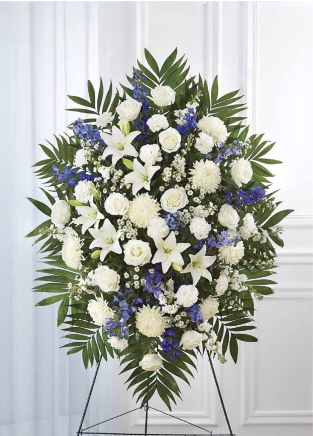 White with Blue Standing Spray - A large, oval standing spray in serene white and cool blue creates a respectful tribute for sympathy or funeral services. This white funeral spray combines lilies, roses, carnations,  so the person you're honoring will be remembered with quiet elegance-the standing easel presentation ensures it reads clearly at the service and from a distance.  Dense clusters of white lilies, white roses and white carnations and chrysanthemums form the luminous center, punctuated by blue delphinium and smaller blue accents for a calming contrast. Broad green palm fronds frame the arrangement, giving a strong, formal outline and clean silhouette appropriate for a memorial setting. The scale is substantial and designed for display on a standing easel; colors are white, blue and green as shown.  For expressing sympathy or offering comfort after a loss, this spray conveys dignity and peace-the kind of floral tribute that helps mourners feel seen. If you need help with delivery timing to a funeral home or special wording for a remembrance card, we can assist when you place your order.