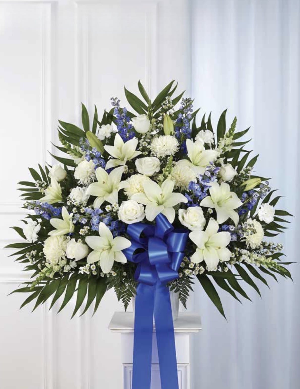 White with Blue Pedestal Arrangement - A serene arrangement in white and blue crafted for sympathy and memorial services, this flower arrangement offers a dignified tribute that honors the person you're remembering. White lilies and white roses take center stage to convey reverence and peace, while clusters of blue blooms and delicate white filler add quiet color and texture-an arrangement designed to provide comfort and a calming visual presence at a service.  Broad, symmetrical, and formal in design, the spray is framed with dark green foliage for contrast and finished with a vivid blue ribbon tied at the base. It's scaled for ceremonial display so it reads clearly in a chapel or funeral setting. This is the kind of arrangement that helps the recipient feel respected and remembered; order with confidence for next-day delivery to most services, and contact us for placement preferences or personalization requests.