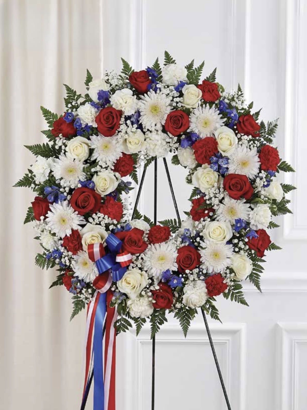 Red, White &amp; Blue Wreath - A large circular sympathy wreath in red, white &amp; blue makes an immediate, respectful statement for funerals, memorials, or Veterans Day remembrances. The formal presentation-mounted on a metal easel stand and tied with a red, white and blue ribbon-signals a tribute meant to honor the person you're remembering, offering a dignified visual that comforts mourners and shows deep respect.  Dense clusters of red and white roses are paired with white chrysanthemums, red carnations, small blue accent flowers, and sprays of baby's breath for a textured, patriotic palette of red, white, blue, and green. The wreath's full, circular shape reads as a complete expression of remembrance; the ribbon adds the finishing ceremonial touch. Sized for display on the included easel, this arrangement is crafted to be seen from a distance and to anchor a service or graveside tribute with grace.