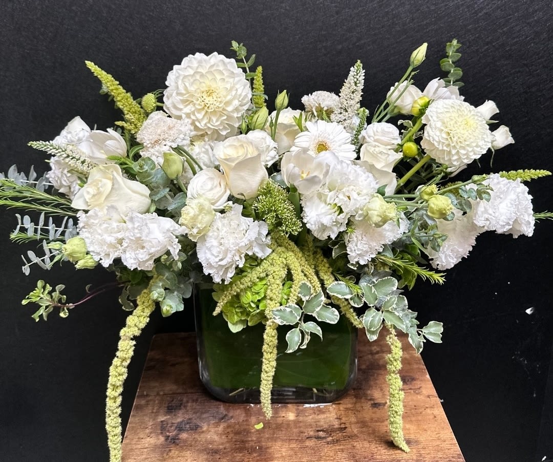 Timeless Grace - Soft white dahlias and garden roses take center stage in this elegant flower arrangement for weddings, sympathy, or any moment that calls for timeless grace. The low-to-medium centerpiece is designed to make the person you're thinking of feel quietly celebrated-serene, seen, and comforted-through a refined palette of whites and soft greens with just a hint of mixed color for warmth.  A layered mix of dahlias, roses, carnations, chrysanthemums, daisies, lisianthus and snapdragons creates a textural composition: large, dahlia-like blooms provide focal weight, airy snapdragons and lisianthus lend vertical movement, and trailing green elements soften the edges. Arranged in a clean square glass vase, the design reads modern yet organic, ideal for a dining table, altar, or bedside surface. Sized as a full but low-profile centerpiece, it's easy to place and enjoy; simple fresh-water care keeps blooms looking their best.