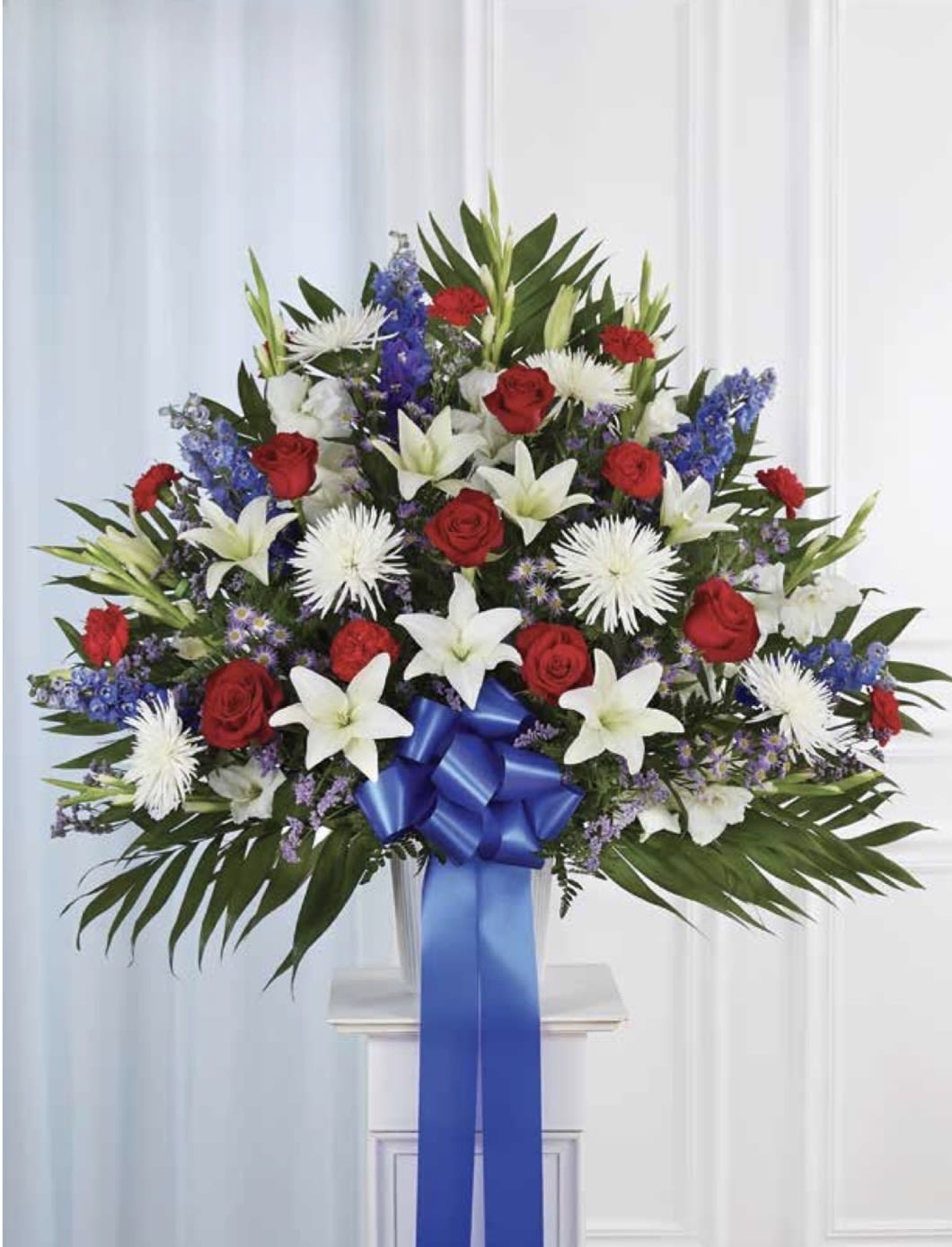 Red, White &amp; Blue Pedestal Arrangement - A formal, ceremonial arrangement crafted in bold patriotic hues-the Red, White &amp; Blue pedestal arrangement is a dignified choice for funeral or memorial services. This flower arrangement combines red roses and white lilies to create a clear red-white-blue statement that honors the memory of a loved one and reads respectfully from the front of a sanctuary or graveside.  Dense and symmetrical, the arrangement layers velvety red roses with large white lilies, blue delphinium accents, other white blooms, and dark green foliage for contrast and depth. A wide blue ribbon and bow cascade from the center, reinforcing the solemn, polished presentation typical of memorial florals.  Perfect for honoring military service, patriotic holidays, or any memorial where you want to express respect and remembrance, this arrangement conveys dignity and heartfelt sentiment. For delivery to a service or cemetery, please confirm date, time, and location when ordering so the pedestal can be set up as requested.