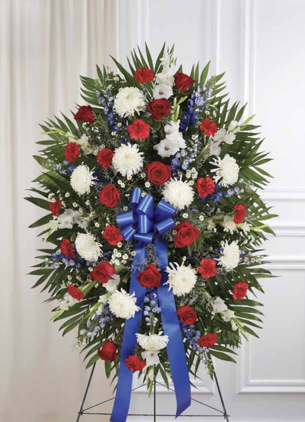 Red, White &amp; Blue Standing Spray - A formal, ceremonial standing spray in red, white &amp; blue crafted for memorial or funeral services-designed to honor and commemorate. The traditional arrangement places red roses and carnations alongside white chrysanthemums with touches of blue delphinium and smaller blue accents, the palette arranged to convey respect and gratitude. A prominent blue ribbon tied near the center and broad palm-style greenery give the spray a dignified, full silhouette ideal for a graveside tribute or service display.  This large spray is mounted on a standing easel for immediate presentation and features a dense, symmetrical composition that reads clearly from a distance. Textures range from the velvety rose petals and ruffled carnations to the crisp, massed chrysanthemums and spiky delphinium, creating visual depth while keeping the design solemn and unified. Choose this arrangement to express patriotic pride, deep sympathy, or heartfelt remembrance-the kind of floral tribute that makes the person being honored feel truly respected.