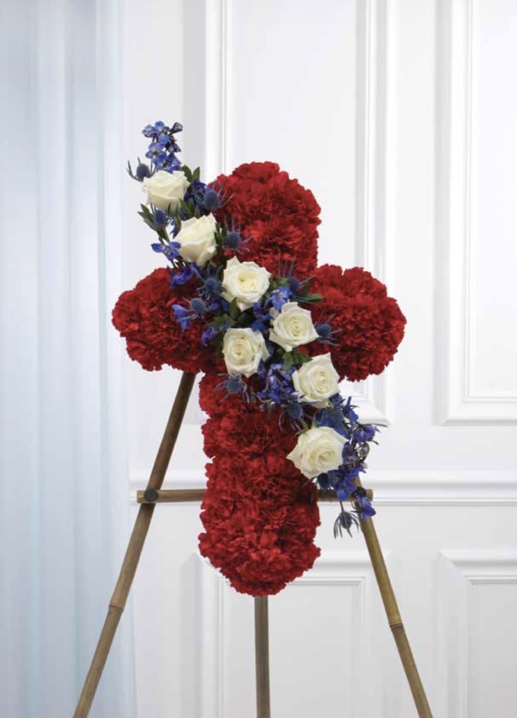Red Cross with a White and Blue Cluster - A dignified memorial cross made from rich red carnations with a diagonal cluster of white roses and small blue accent flowers-this funeral arrangement brings a traditional, ceremonial presence to services and graveside tributes. Designed as a flower arrangement for memorials and funerals, the red cross shape conveys lasting respect while the white and blue diagonal cluster adds contrast and a quiet, comforting focal point.  The cross is densely covered in red carnations for a full, textured surface; a band of white roses and delicate blue filler flowers sweeps across the center to create a stately accent. The piece is mounted on an easel stand for upright display at services or remembrance settings, and its proportions present clearly against a light interior or outdoor backdrop. This arrangement offers a solemn, heartfelt way to honor someone's memory-sized and styled for traditional services and simple to place at the front of a ceremony.