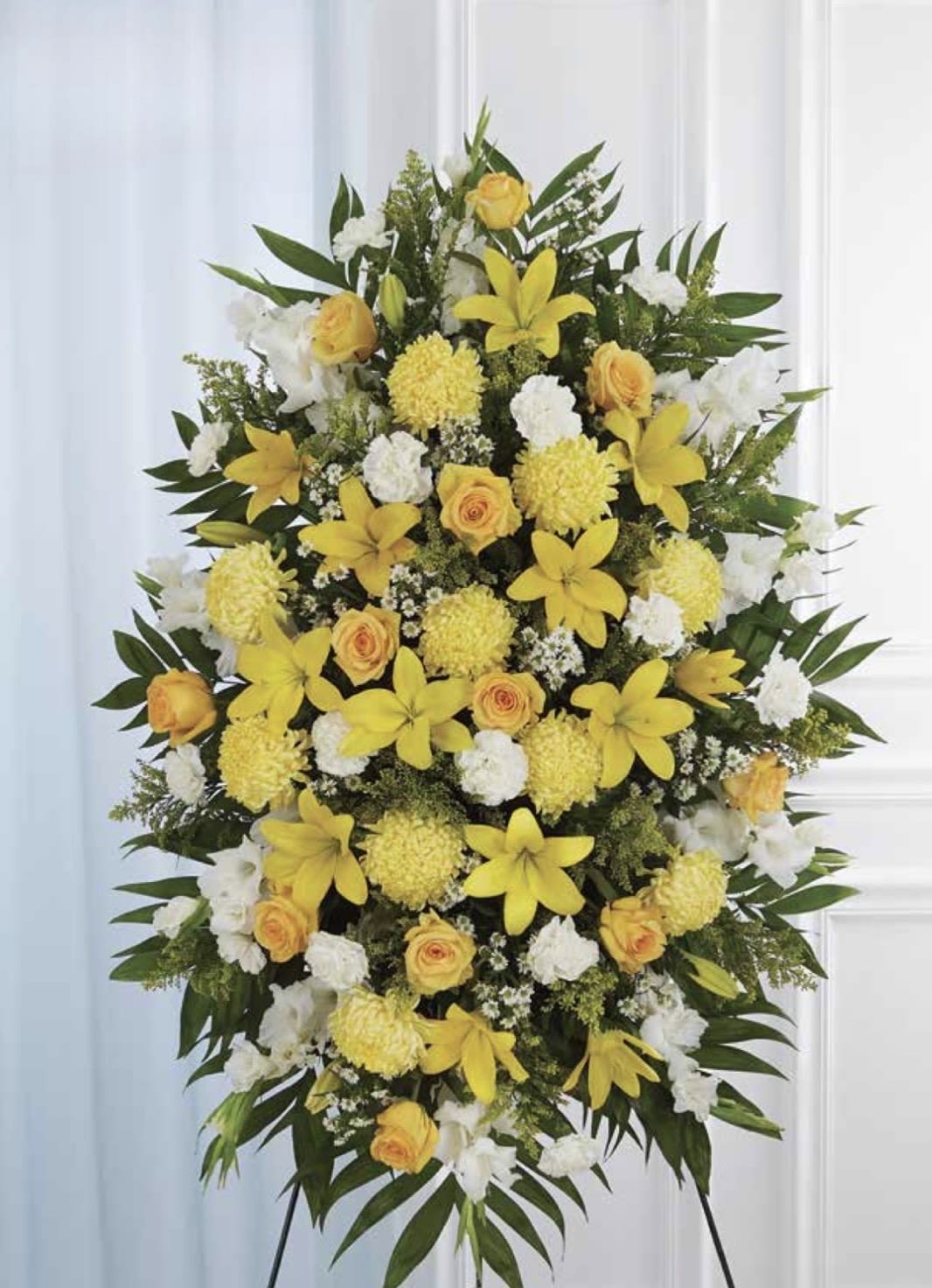 White and Yellow Standing Spray - A tall, dignified standing spray that speaks with quiet brightness-perfect as a sympathy arrangement or memorial tribute. The white and yellow standing spray places yellow roses and open lilies beside pristine white carnations and round yellow chrysanthemums, creating a warm, uplifting palette that honors a life while conveying comfort. Its formal, oval shape and balanced symmetry make it appropriate for funeral services and graveside ceremonies.  Dense layers of dark green foliage cradle each bloom so the colors read clearly from a distance; the texture shifts from the soft ruffles of carnations to the sculpted petals of lilies and the classic form of roses. Mounted on a sturdy easel, the spray is designed to stand tall and visible during services. Choose this arrangement when you want the person you're remembering to feel acknowledged and celebrated-size and presentation are intended for display at memorial services, and standard care (keep blooms cool and sheltered from direct sun or wind) will help maintain its fresh appearance through the event.