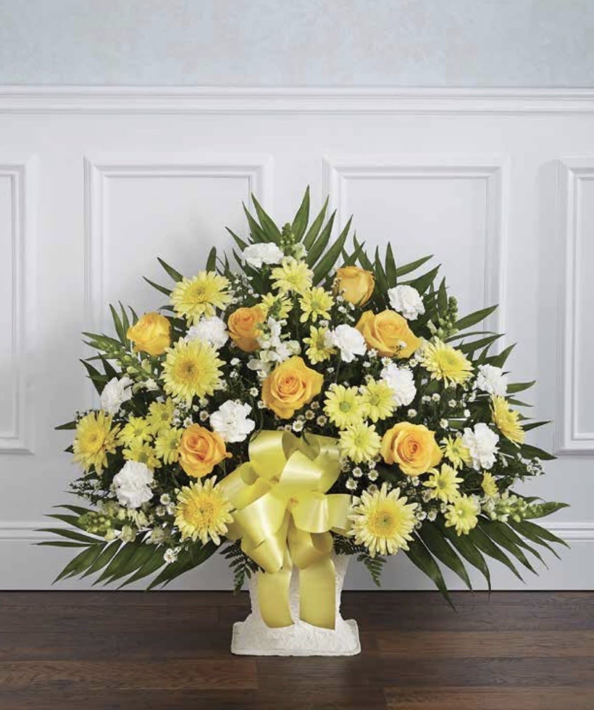 White and Yellow Pedestal Arrangement - Sunny yellow roses and pale daisy-like blooms create an uplifting focal point in this white and yellow pedestal arrangement-perfect as a cheerful sympathy spray, celebration piece for congratulations, or a thoughtful thank-you. The arrangement's rounded spray and formal pedestal silhouette make an immediate bright statement so the person you're thinking of will feel noticed and comforted by the warm, optimistic colors.  A mix of yellow roses, pale yellow daisy-style flowers, white accent flowers and carnations are gathered atop a white pedestal vase, with long pointed palm fronds fanning out behind to add layered texture and height. A wide yellow ribbon tied at the front of the vase provides a soft, decorative finish; overall tones are yellow, white, and green for a clean, sunny palette that reads both joyful and respectful.  Ideal for sending to honor a milestone, celebrate a life, or simply lift someone's spirits, this piece arrives ready to display on an entry table or at a service. The pedestal presentation gives it presence without extra assembly; keep blooms in fresh water, out of direct sun, and recut stems every few days to extend vase life.