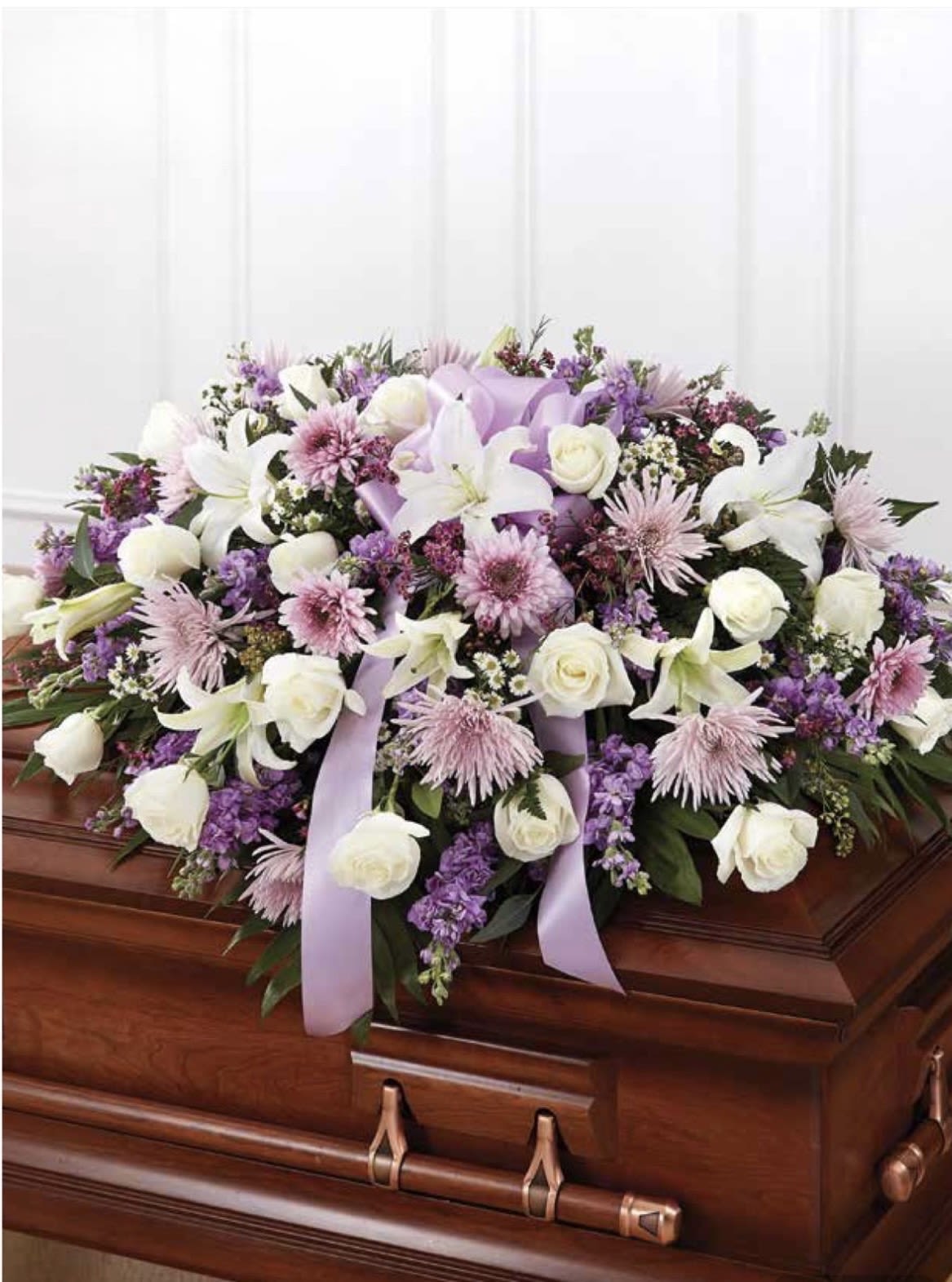 White and Lavender Casket Spray - Soft white lilies and roses mingle with pale lavender chrysanthemums and clusters of purple accents in this white and lavender casket spray, designed for sympathy and memorial services. The arrangement's low, full profile lays gracefully across a wooden casket, creating a serene, formal tribute that quietly honors the person you're remembering.  Dense dark-green foliage anchors the flowers while pale lavender ribbon is artfully draped through the design for a restrained, elegant touch. The palette of white, purple, and green keeps the look gentle and respectful; proportions are made to cover the casket top with a symmetrical, balanced silhouette that reads well from the front and sides.  Made to express condolences and respect, this spray is the kind of tribute that helps loved ones feel comforted and seen during services and visitations. For delivery and placement on the wooden casket, please indicate service time and any personalization requests when ordering so we can ensure a timely, dignified presentation.