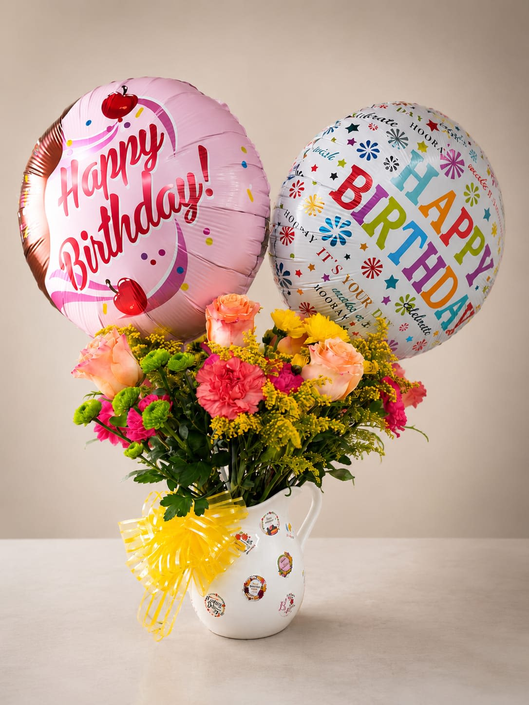 Radiant Birthda - A playful cluster of pink roses, carnations, and bright yellow filler blooms topped with two large birthday foil balloons makes this flower arrangement an unmistakable birthday bouquet that will make the person you're celebrating feel cheered and seen. The mixed-color palette-soft pinks, sunny yellows and touches of white-reads joyful and festive, perfect for birthdays, congratulations, or any upbeat milestone.  The flowers are arranged compactly in a white ceramic pitcher-style vase decorated with small birthday stickers and finished with a sunny yellow ribbon bow, giving the gift a charming, ready-to-display look. Textures move from the velvety rose petals to the ruffled carnations and crisp chrysanthemum accents, creating visual depth and long-lasting appeal. Two large foil balloons rise above the arrangement for immediate party impact.  Send this when you want them to feel truly celebrated-ideal for a friend, family member, or colleague marking a birthday or small victory. The pitcher vase arrives ready to display; refresh stems with water as needed to extend enjoyment and keep blooms looking fresh.