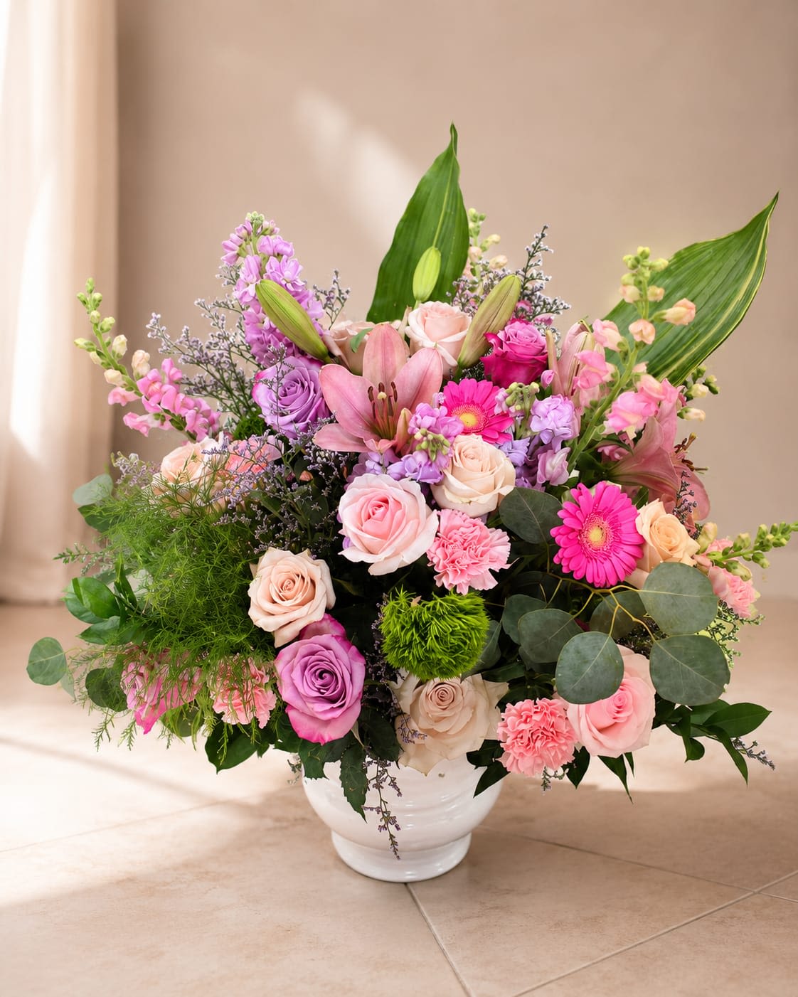Blushing Beauty - A lush, pastel-hued bouquet that reads like a love letter in flowers-Blushing Beauty is a large birthday bouquet and anniversary arrangement designed to make her feel truly celebrated. Soft pink roses and blush lilies set a romantic tone while lavender and white accents add a gentle, sophisticated contrast; the overall impression is feminine and uplifting, perfect for Mother's Day or to mark a special milestone.  This rounded design is arranged in a white ceramic vase, creating a clean, elegant base for the abundant mix of roses, lilies, gerbera daisies, carnations and tall spires of mixed accent flowers. Broad decorative leaves and airy greenery add height and structure, while vivid magenta touches bring moments of bright, joyful color through the soft palette. The composition is generous in scale and texture-petal-rich blooms, open lilies, and sculptural foliage combine for long-lasting visual impact.  Give this to the person you're thinking of when you want them to feel cherished and seen; it communicates affection, admiration, and celebration in equal measure. The white ceramic vase makes it ready to display on arrival-simply trim stems and refresh water as needed to extend vase life.