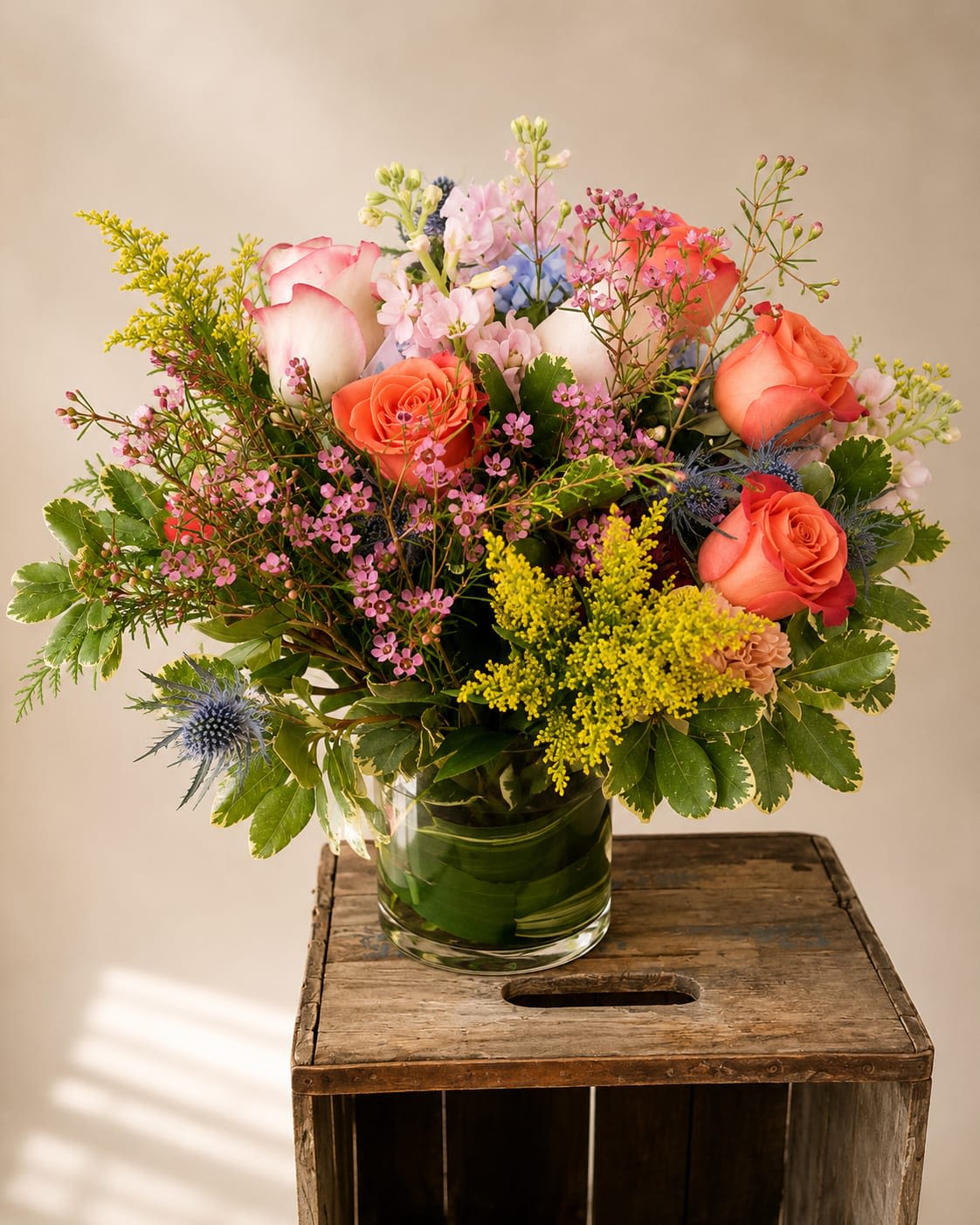 Sunset Bloom - A full, rounded bouquet of coral and blush roses that reads like a painted sunset-perfect as a birthday bouquet, anniversary arrangement, or an everyday gift to brighten someone's day. The mix of warm orange and soft pink roses with pops of yellow and pale blue filler flowers creates a cheerful, romantic impression the person you're thinking of will notice the moment it arrives.  This flower arrangement pairs dense, velvety rose heads with airy clusters of small pink, yellow, and pale blue blooms and branching green accents that give height and gentle movement above the main bloom line. The palette of pink, orange, yellow, and green feels fresh and springlike, and the stems are gathered in a clear glass vase that shows off the natural lines and adds a clean, contemporary touch.  Choose this arrangement when you want someone to feel celebrated and seen-whether it's for a birthday, an anniversary, or simply because you're thinking of them. Standard sizing fits comfortably in the included clear glass vase; blooms should be kept in fresh water and trimmed a little every few days to extend vase life.