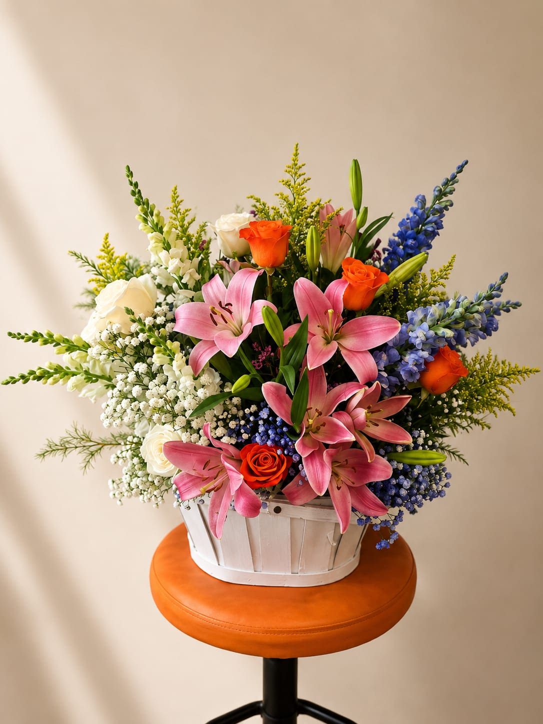 Spring Radiance - Large pink lilies take center stage in this Spring Radiance flower arrangement, making it a joyful choice for a birthday bouquet, congratulations gift, or bright everyday surprise. The mix of pink, white, orange and blue blooms reads cheerful and celebratory-the kind of arrangement that will make the person you're thinking of feel genuinely seen and celebrated.  This full, rounded composition pairs large pink lilies with orange and white roses, tall white snapdragon spikes, cheerful daisies, and clusters of small white filler blooms for texture. Blue accent flowers punctuate the palette, adding lively contrast and depth. The flowers are arranged in a white basket that gives a rustic, hand-delivered look and lets blooms extend outward in several directions for a lush, abundant effect.  Perfect for sending congratulations, brightening a bedside table, or marking a special birthday, Spring Radiance arrives ready to display. For best longevity, keep stems in fresh water, trim stems at an angle before arranging, and place away from direct heat or sunlight to help the recipient enjoy the vibrant colors longer.