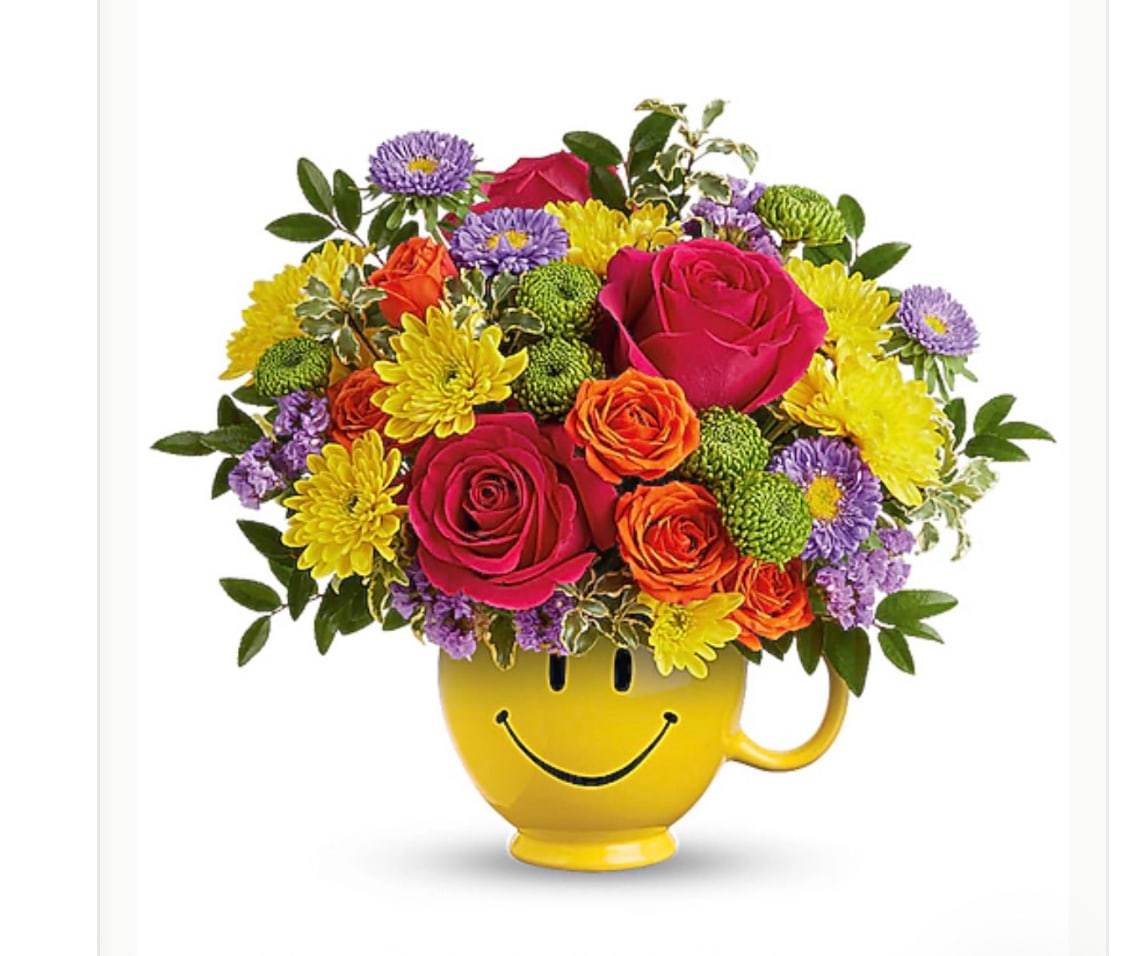 HAPPY BOUQUET - Bright, playful, and impossible to ignore, this happy bouquet is a birthday-ready flower arrangement that also works beautifully for just-because, spring and summer celebrations. A glossy yellow ceramic mug with a cheerful smiley-face graphic anchors the design, so the person you're thinking of gets both fresh blooms and a fun keepsake they can reuse. The sunny, casual vibe makes it an upbeat pick for Labor Day barbecues or a small Memorial Day remembrance that feels warm rather than formal.  Densely packed above the mug, vivid pink roses and bright orange spray roses mix with sunny yellow chrysanthemums and small purple aster-style daisies for a lively, multi-colored display. The compact, rounded silhouette keeps the arrangement modern and gift-ready, with green foliage tucked in to add texture and a fresh contrast to the warm and cool color palette. The mug adds practical value and a pop of color that amplifies the bouquet's cheerful statement.  Perfect for the person who deserves a smile-whether marking a birthday, saying thank you, or brightening an ordinary day-this arrangement arrives ready to display on a kitchen counter, desk, or bedside table. Standard care: keep flowers in fresh water, trim stems occasionally, and avoid direct midday sun to extend vase life; the ceramic mug can be washed and reused after flowers are removed.