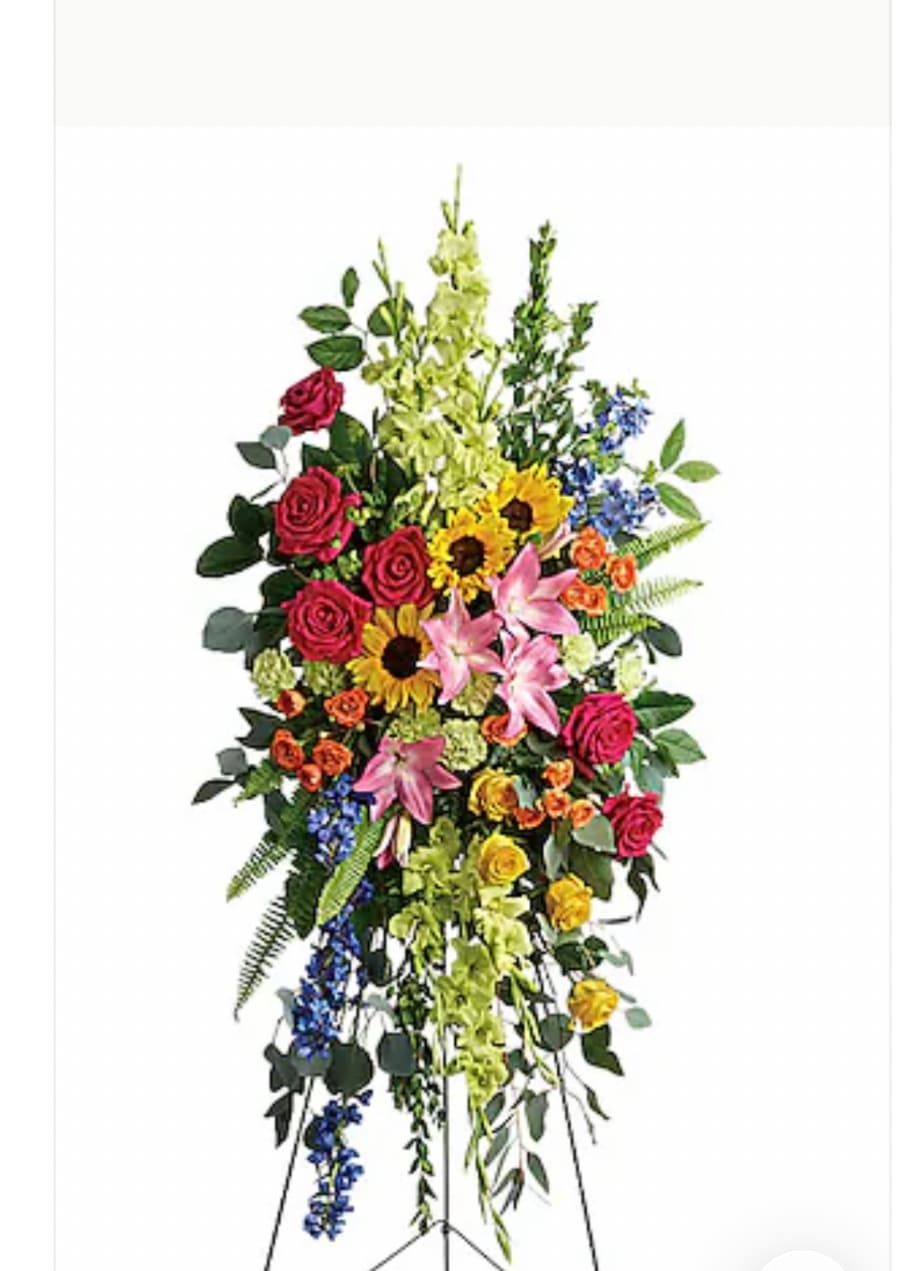 LOVE LIVES ON SPRAY - A tall standing spray that balances warmth and reverence, this sympathy spray features pink roses and pink lilies paired with sunny yellow sunflowers to create a hopeful, consoling presence for a memorial service or celebration of life. The vertical, easel-mounted design reads as formal and respectful while the lively palette of pink, yellow, purple and fresh green keeps the tribute feeling personal-perfect for the person you're honoring when you want their memory to feel bright and cherished.  Dense clusters of roses, lilies, sunflowers, snapdragons and small blue accent blooms form a layered, cascading composition with long trailing elements and mixed greenery for a full, elegant silhouette. Mounted on a standing easel, it offers strong visual impact at the front of a service or beside a casket; the varied textures-from the sunflower heads to the soft rose petals and spiky snapdragons-give depth and movement across the piece.  Choose this spray when you want to express sympathy while celebrating a vibrant life: it reads as comforting to grieving family members and uplifting to those sharing memories. The easel display is delivered ready to place; for best appearance, keep flowers in a cool location away from direct heat and confirm delivery timing with the service planner so the arrangement arrives fresh and properly set.