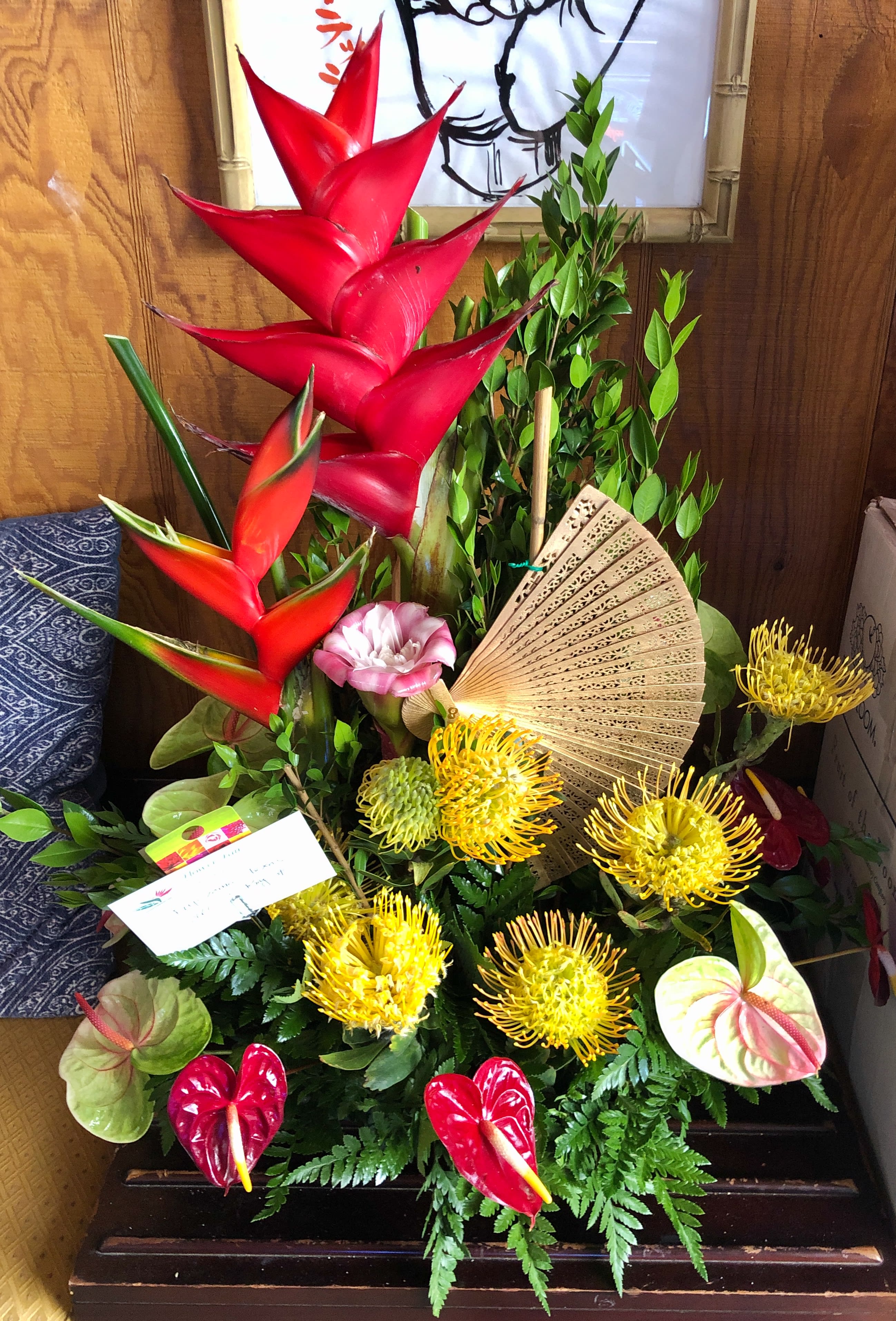 Tropical flower arrangement with red heliconia, yellow pincushion blooms, and an open fan