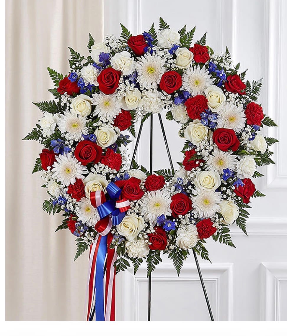 PATRIOTIC PASSION WREATH - A large heart-shaped memorial wreath in red, white and blue that reads as a patriotic tribute for a funeral or remembrance service. This funeral wreath blends roses, carnations, daisies and delicate sprays of baby's breath into a dense, layered heart that makes a solemn, respectful statement - the kind of sympathy arrangement that lets the person you're honoring feel remembered and respected.  The wreath is finished with small blue accent flowers and a red-white-and-blue ribbon at the lower left, and it arrives mounted on a black easel for immediate chapel or graveside display. Textures range from the satin petals of roses to the ruffled carnations and the airy baby's breath, creating depth and a comforting visual rhythm in traditional red, white, blue and green tones.  Designed specifically for memorial services, patriotic holidays, or to honor a veteran, this heart-shaped tribute communicates reverence and gratitude. Easy to place on site with the included easel and ribbon, it's sized to be a prominent focal piece while remaining tasteful and ceremonial.