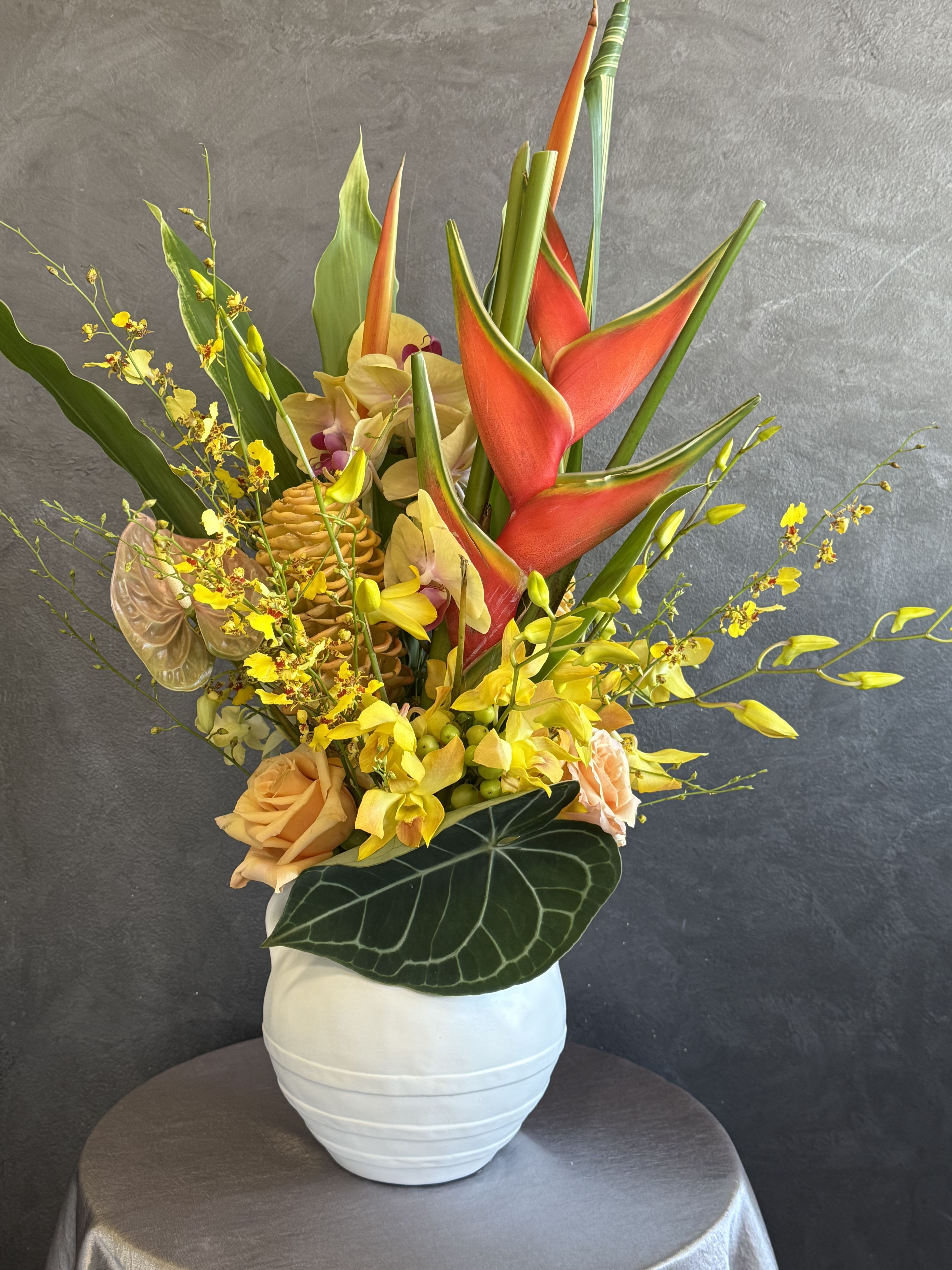 Tropical arrangement with red heliconia, yellow orchids, and peach roses in a white vase