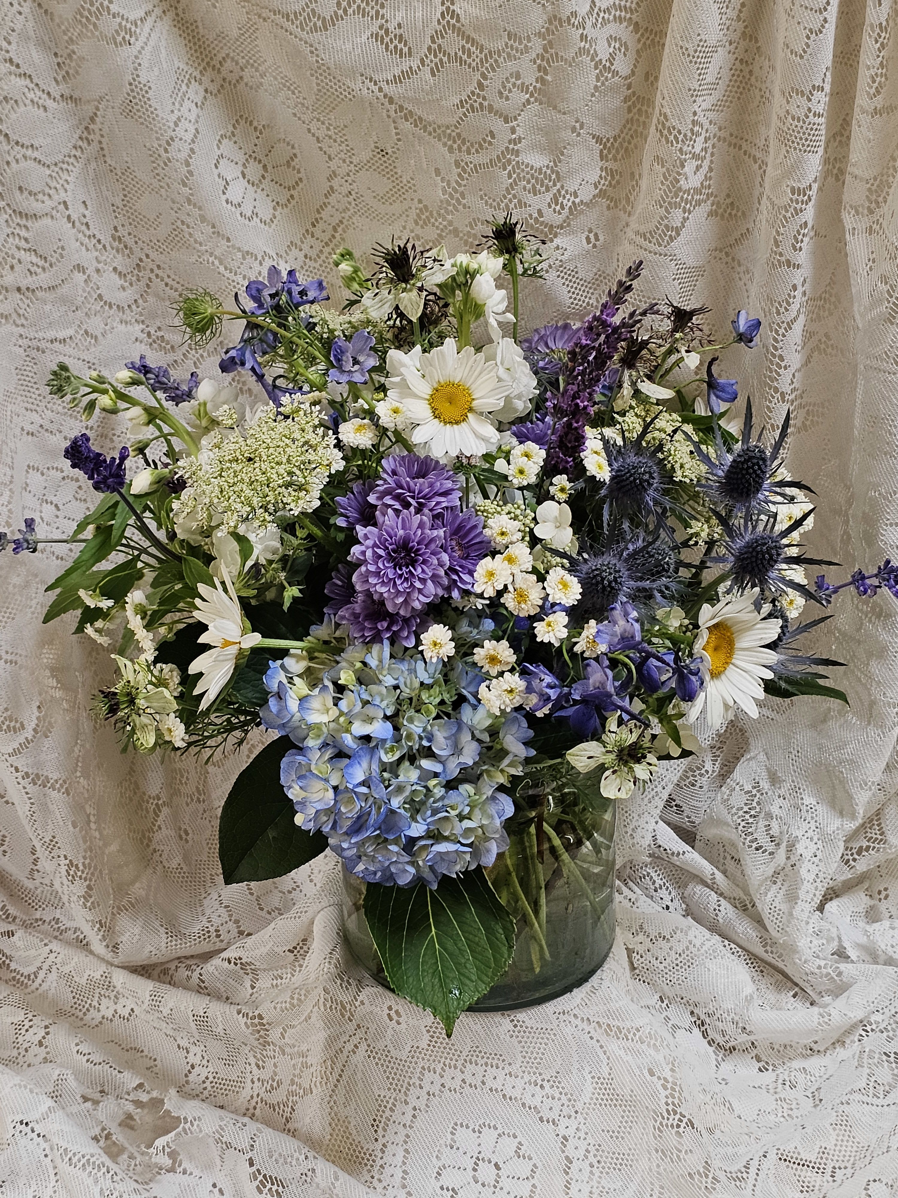 Blueberry Cobbler - Sweet, fresh, and overflowing with summer charm, Blueberry Cobbler is a lush, plump floral arrangement inspired by the season's favorite dessert. Designed in a low vase, this abundant centerpiece features a refreshing palette of soft whites and rich, summery blues, creating a look that feels both elegant and effortlessly inviting. Blueberry Cobbler brings the fresh, carefree spirit of summer indoors.
