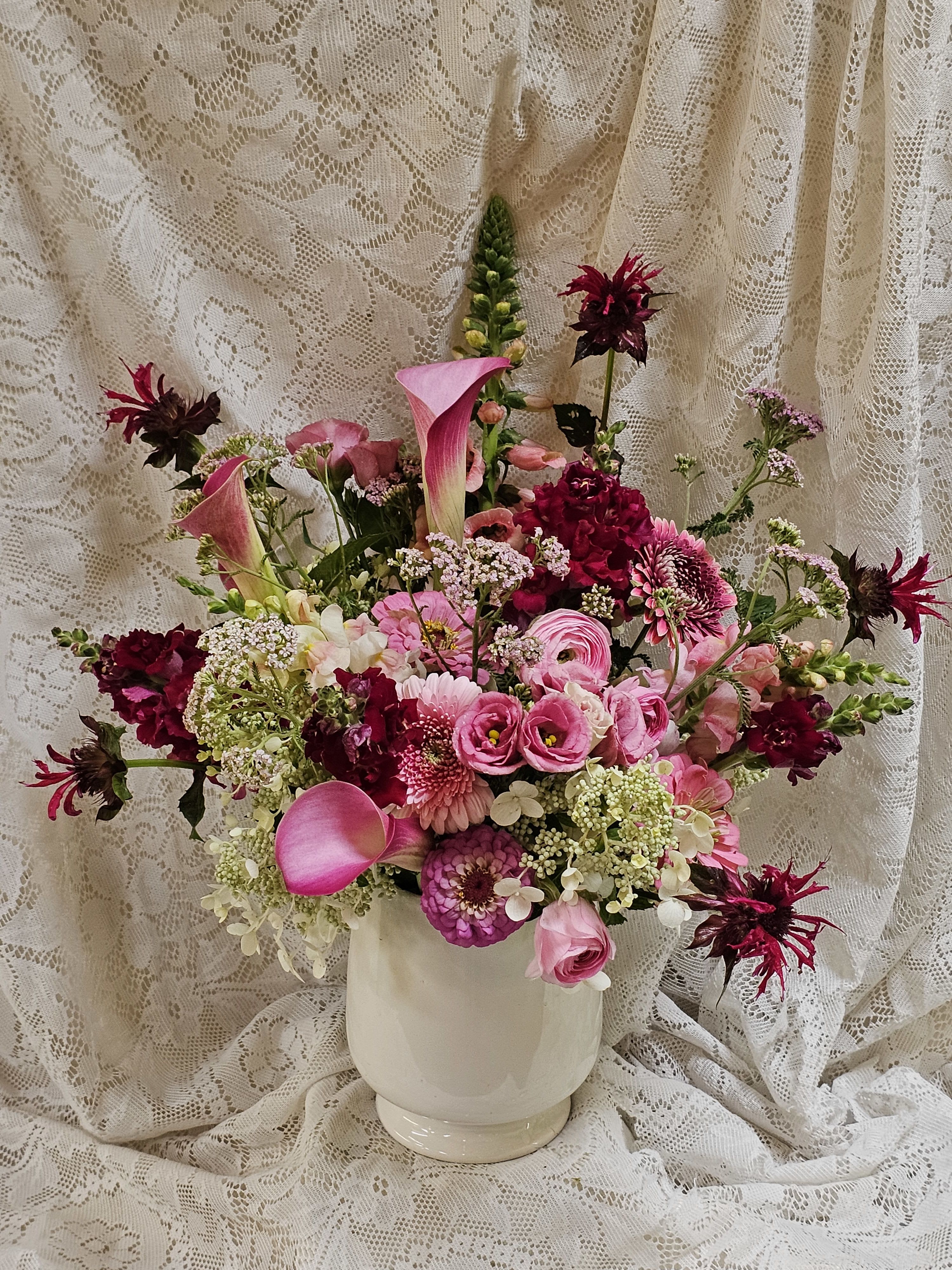 Black Raspberry - Rich, velvety, and irresistibly beautiful, Black Raspberry is a lush floral arrangement inspired by the decadent colors of its namesake ice cream. Designed in a timeless white ceramic vase, this abundant, garden-style arrangement features layers of deep raspberry, plum, burgundy, and blackberry tone blooms, beautifully balanced with soft blush and creamy accents that evoke ribbons of sweet vanilla.  Overflowing with premium flowers and luxurious texture, Black Raspberry is full of depth, dimension, and romance. The crisp white ceramic vase perfectly complements the rich color palette, allowing every bloom to shine while adding a clean, modern touch. Bold yet elegant, this arrangement makes a stunning gift or centerpiece for any occasion.