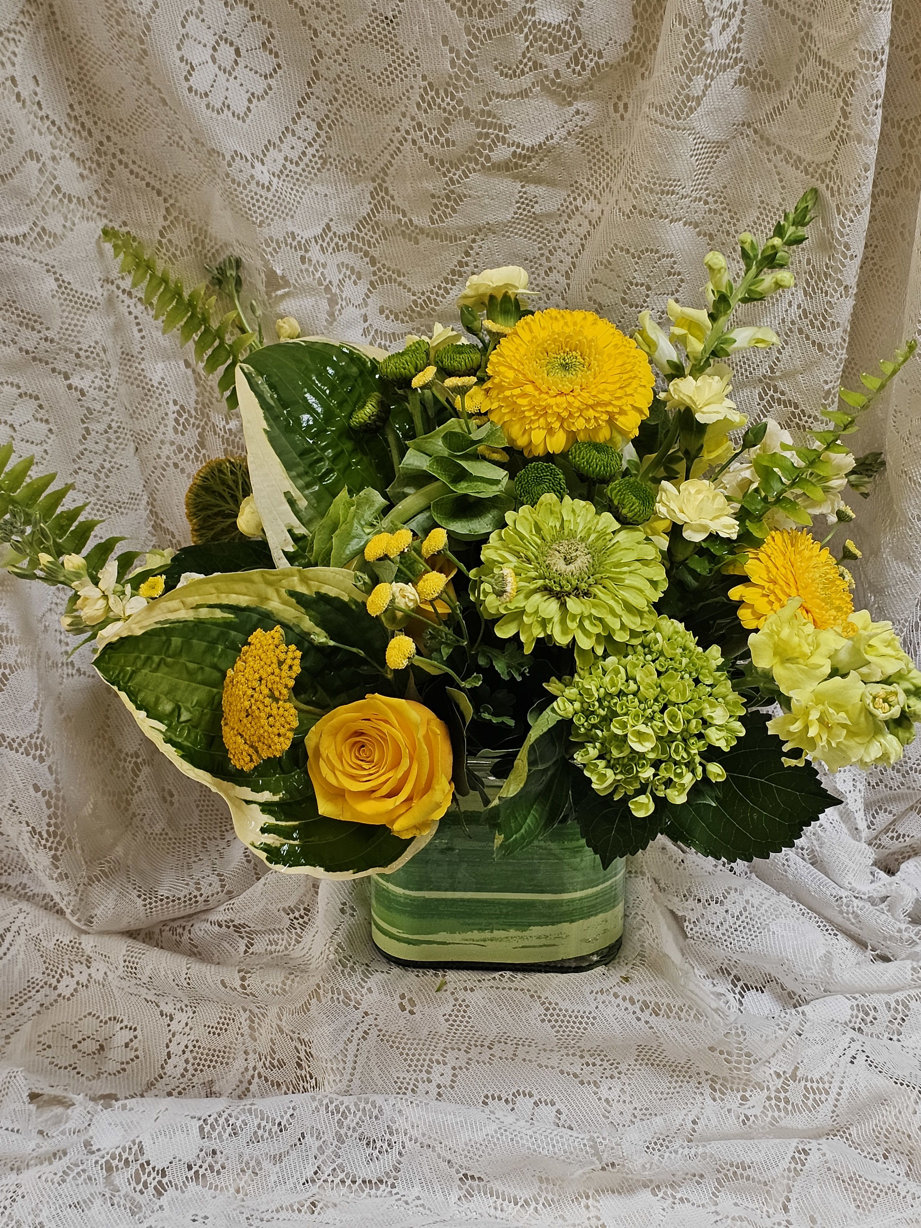 Key Lime Pie - Fresh, zesty, and bursting with summer color, Key Lime Pie is a lush floral arrangement inspired by the bright, refreshing flavors of the classic dessert. Designed in a low glass vase with a leaf wrap, this vibrant arrangement features cheerful lime greens, sunny yellows, and creamy blooms for a look that's crisp, clean, and full of seasonal charm. Garden-fresh zinnias bring playful texture and pops of sunshine, while lush hosta foliage creates a naturally elegant foundation that enhances the arrangement's vibrant color palette.