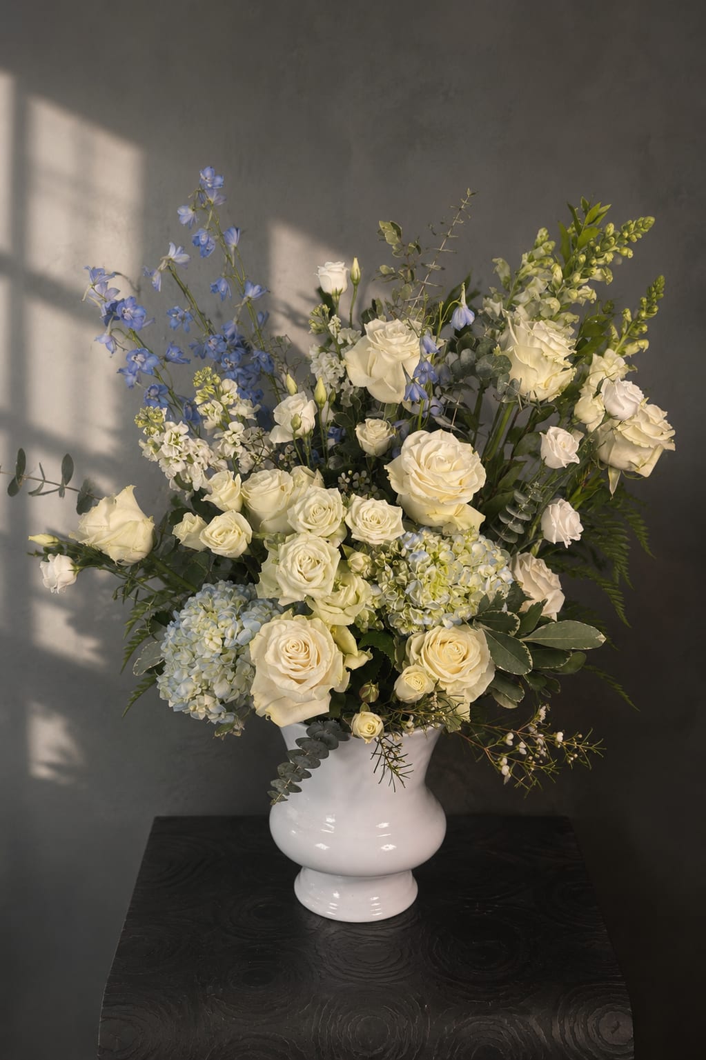 Silent Comfort - When words are not enough, Silent Comfort speaks through grace and quiet beauty.  This elegant sympathy arrangement features creamy white roses, airy blue delphinium, soft blue hydrangea, lisianthus, snapdragons, and seasonal accents, thoughtfully arranged in a timeless white ceramic vase. The soothing palette of white and blue symbolizes peace, remembrance, and hope, creating a meaningful tribute to honor a cherished life.  Designed to bring comfort during difficult moments, Silent Comfort is a heartfelt gesture for funeral services, memorial gatherings, or to express condolences to family and loved ones. Each arrangement is handcrafted with care by our florists, ensuring every tribute is as unique as the life it celebrates.