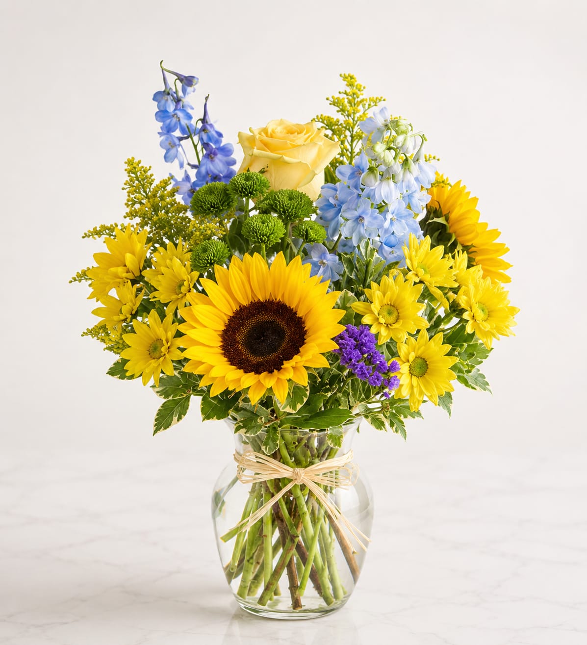 Golden Sunshine Garden™ - Brighten any day with the cheerful beauty of Golden Sunshine Garden™. This vibrant arrangement features radiant sunflowers, a classic yellow rose, blue delphinium, yellow daisy mums, green button poms, purple statice, golden solidago, and premium seasonal greenery, all beautifully arranged in a clear glass vase with a rustic raffia bow. A joyful gift for birthdays, congratulations, thank-you wishes, get well sentiments, or simply bringing sunshine into someone's home.