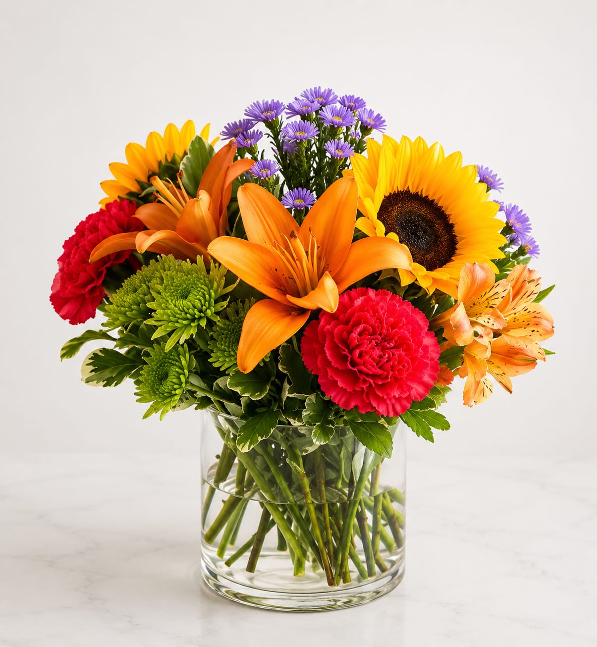 Sunset Celebration Bouquet™ - Bursting with warmth and vibrant color, Sunset Celebration Bouquet™ is a stunning arrangement featuring radiant orange lilies, hot pink and red roses, cheerful sunflowers, bright carnations, lavender stock, green button poms, alstroemeria, and fresh seasonal greenery, all beautifully arranged in a clear glass vase. This eye-catching bouquet is perfect for birthdays, celebrations, congratulations, thank-you gifts, or adding a splash of sunshine to any occasion.