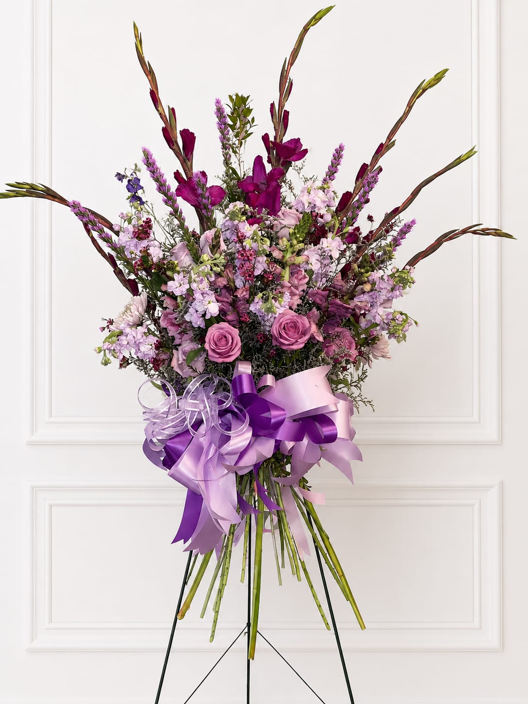 Peaceful Reflections Standing Spray - A tall, sculptural standing spray that greets the room with layered pink and purple blooms-this sympathy and funeral spray makes a comforting, romantic statement for a memorial service or celebration of life. The arrangement's dramatic height and arching stems create a graceful silhouette that stands above the casket or at the front of the sanctuary, offering a visible tribute that feels both reverent and warmly expressive.  The center is a dense, rounded cluster of roses, stock, and other seasonal flowers in soft pinks, purples, whites, and greens, while long snapdragon stems and foliage fan upward to form a sculptural crown. A large, layered lavender and deep purple ribbon bow finishes the piece and the arrangement arrives ready on a floral stand for immediate display. Ideal when you want the recipient's family to feel comforted and honored, this spray is sized for ceremonial impact and designed for easy placement at services.