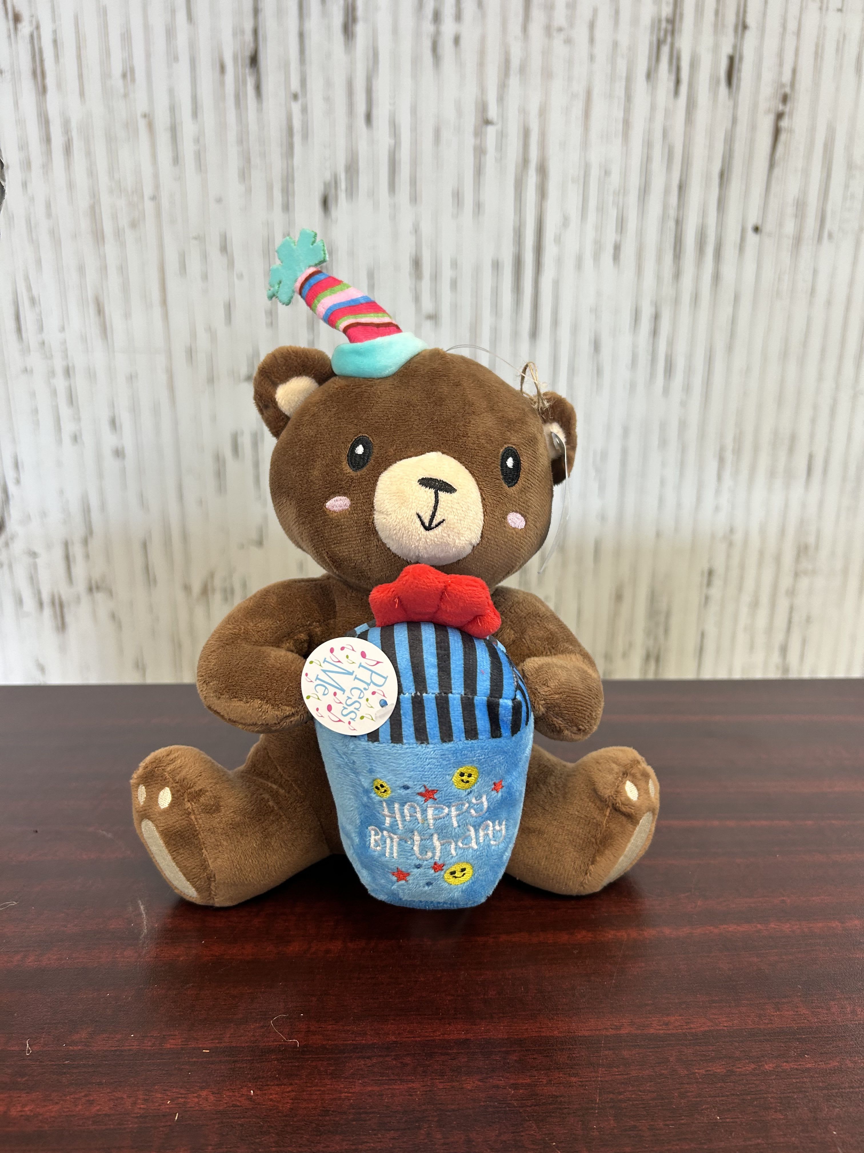Birthday Musical Teddy Bear - A seated brown teddy bear plush holding a blue birthday cupcake makes this birthday arrangement delightfully playful-the perfect birthday gift or party table accent to make someone feel celebrated. The piece reads as a casual birthday bouquet alternative or cheerful add-on gift for kids, friends, or coworkers who prefer whimsical charm over traditional roses.  The bear wears a striped party hat and a bright red bow tie, and the embroidered "Happy Birthday" on the cupcake plush plus small decorative accents reinforce the festive theme. Pink and blue touches give a soft, friendly palette that pairs well with colorful bouquets or gift bags; a hanging tag is included for your message. Sized to sit on a mantel or bedside table, it's an easy, low-maintenance way to send birthday wishes that will be kept and enjoyed long after the day.