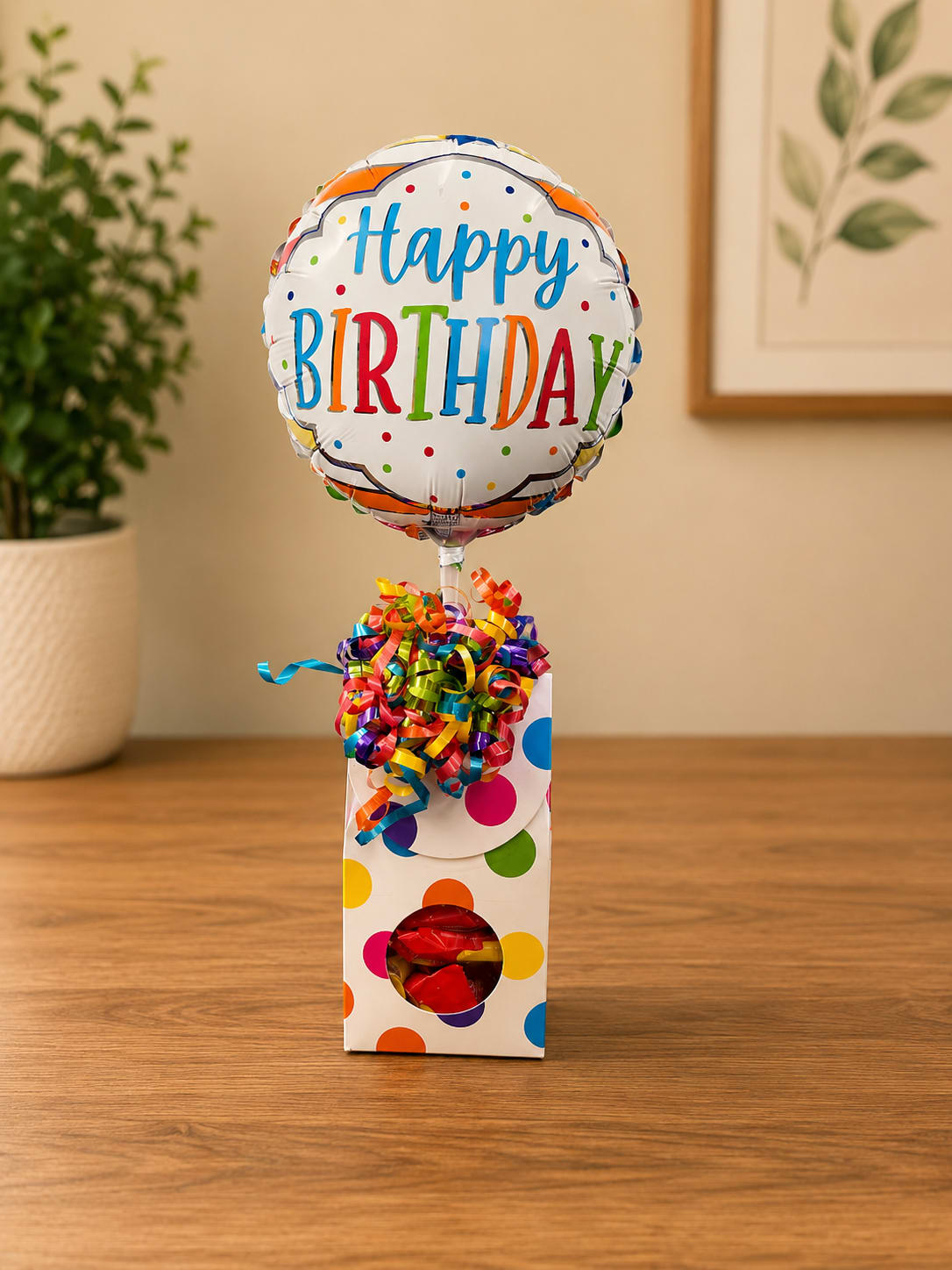 Birthday Candy Gift Box - A round "Happy Birthday" foil balloon bursts with color at the heart of this cheerful birthday gift box, making it instantly recognizable as a festive birthday arrangement. The playful multicolored lettering and confetti dots set a celebratory tone, perfect for birthdays or any upbeat everyday celebration where you want the recipient to feel seen and celebrated.  The balloon is anchored in a white polka-dot gift box filled with visible candies and finished with a dense cluster of curled rainbow ribbons for bright texture and movement. Colors include mixed brights with white accents plus notes of blue and red from the packaging and candies; hard goods are the foil balloon, the decorated gift box, assorted candy, and curling ribbon. Sized to be a compact, hand-deliverable surprise, it arrives ready to brighten a table or desk without extra assembly.  This is a ready-made choice for friends, coworkers, or family when you want to send joy and a sweet treat together-the kind of gift that makes someone smile the moment it's opened. Order with same-week pickup or local delivery to ensure the balloon arrives fully inflated and the candy stays fresh.