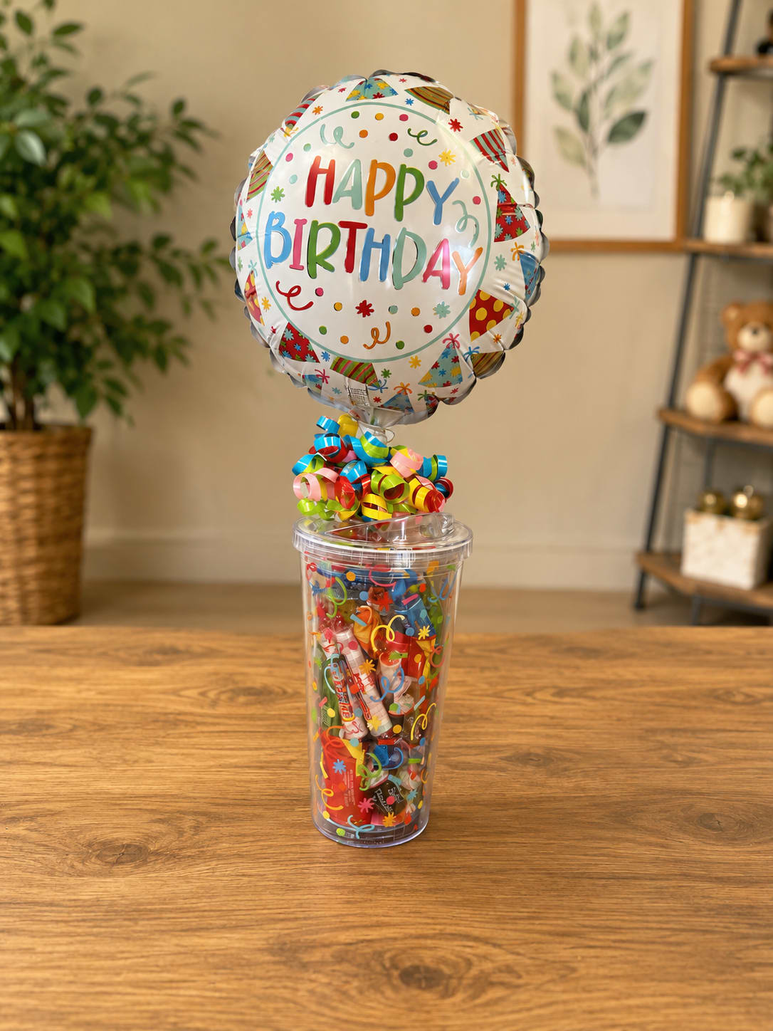 Birthday Celebration Candy Tumbler - A bright "Happy Birthday" foil balloon crowned with curled ribbons makes this birthday bouquet-style gift feel instantly festive - perfect as a birthday centerpiece or a cheerful desk surprise. The Birthday Celebration Candy Tumbler is a playful birthday gift that reads as a celebratory novelty and will make the person you're thinking of smile the moment it arrives.  The balloon is anchored in a clear plastic cup filled with assorted gift filler and confetti-like decorations in mixed colors (white, pink, blue and rainbow accents), creating a compact upright display that fits easily on a table or counter. Curling ribbon adds height and movement while the cheerful printed message keeps the sentiment unmistakable. Ideal for coworkers, friends, or anyone who likes fun, low-maintenance surprises; no watering required and sized to sit wherever the birthday moment will be enjoyed.