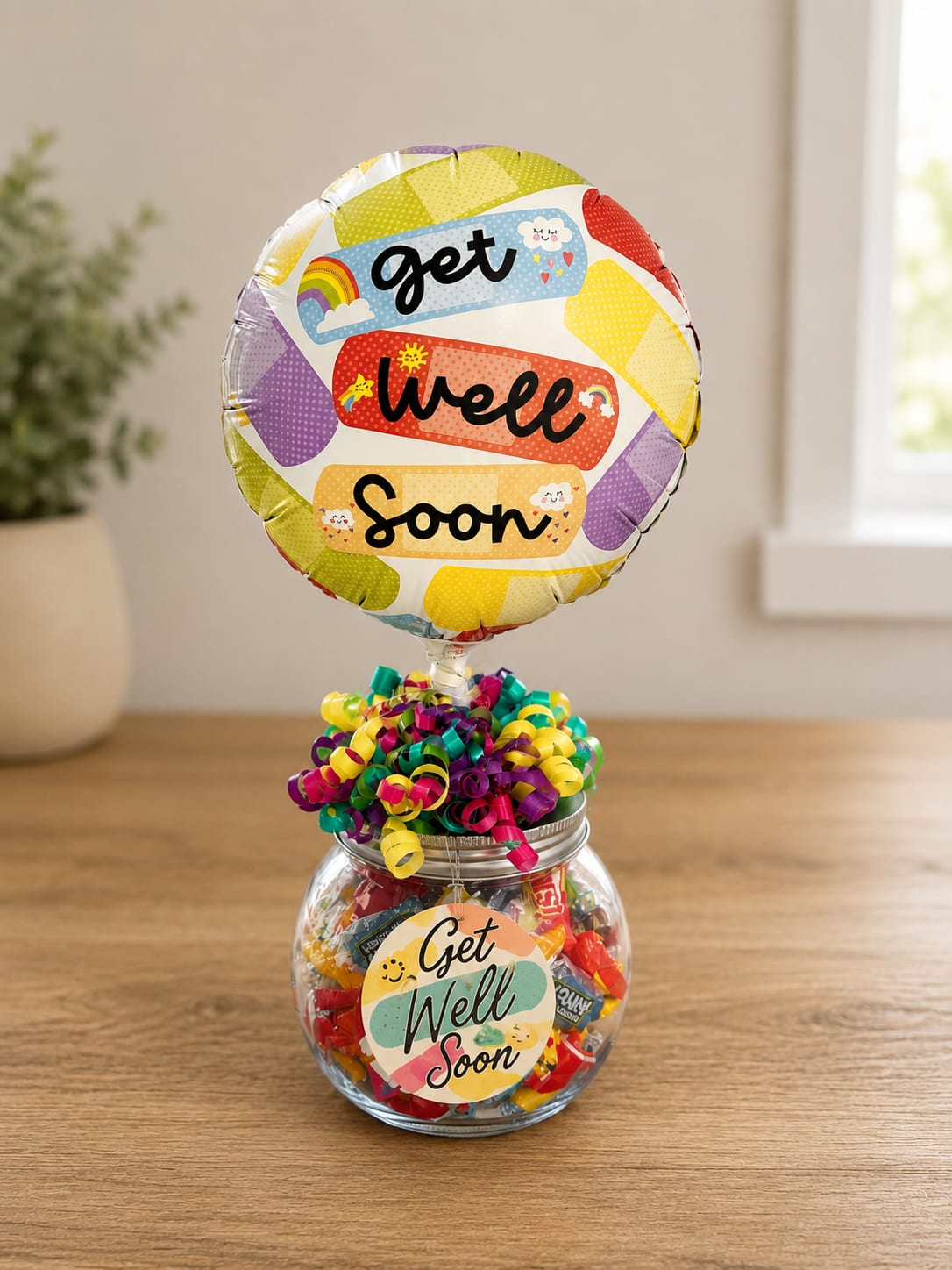 Get Well Candy Jar - Bright, playful, and undeniably cheerful - this get-well gift set presents a round "Get Well Soon" foil balloon paired with a clear glass jar overflowing with assorted wrapped candies, perfect as a get-well bouquet alternative for someone needing a little lift. As a recovery gift or hospital visit present, it reads as lighthearted and comforting: the balloon sends the message while the colorful treats and curling ribbons bring a smile the moment they're seen.  The jar is filled with mixed-color candy in yellow, pink, purple, teal, and red tones and topped with a cluster of bright curling ribbon that links the candy jar to the balloon for a cohesive, celebratory look. Delivered ready to gift with the foil balloon attached, this compact arrangement is easy to carry into a hospital room or set on a bedside table. It's ideal for friends, coworkers, or family members who'd appreciate a sweet, upbeat reminder that you're thinking of them during recovery.