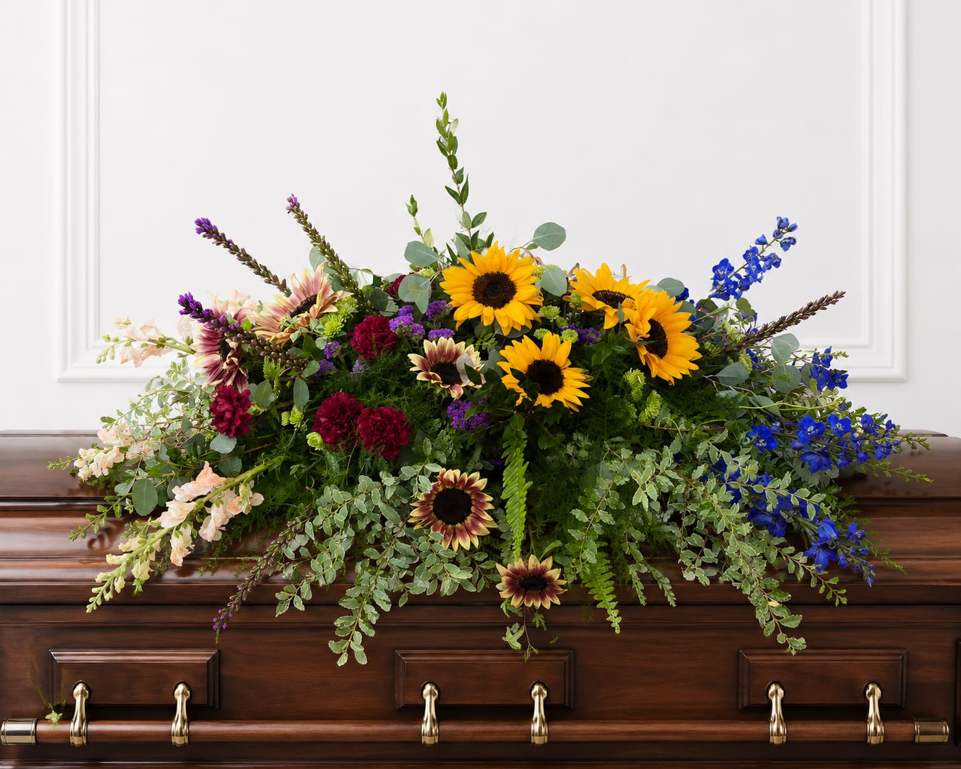 Resting in Grace Casket Spray - Sunflowers anchored at the center give this large casket spray a warm, hopeful focal point - the kind of sympathy arrangement that reads as heartfelt and life-affirming at a funeral or memorial service. Designed as a broad, low spray to lie across a polished wooden casket, its mixed-color palette of yellow, blue, purple and pink creates an expressive, garden-like statement that comforts and honors the person you're remembering.  The spray features bright sunflowers, tall blue delphinium, soft carnations, snapdragons and other mixed blooms in shades of purple, burgundy and blush, all woven with abundant greenery for texture and a natural cascade across the casket. The composition is full and dramatic without being fussy; the varied heights of delphinium and snapdragons give vertical movement while the sunflowers and clustered carnations provide a steady visual center.  Made to convey sympathy with dignity, this arrangement is ideal for family members or close friends seeking a meaningful, visually striking tribute. Sized to cover a standard casket, it arrives ready to place on the polished wooden casket surface; minimal care is required beyond standard floral water and cool-room placement before the service.