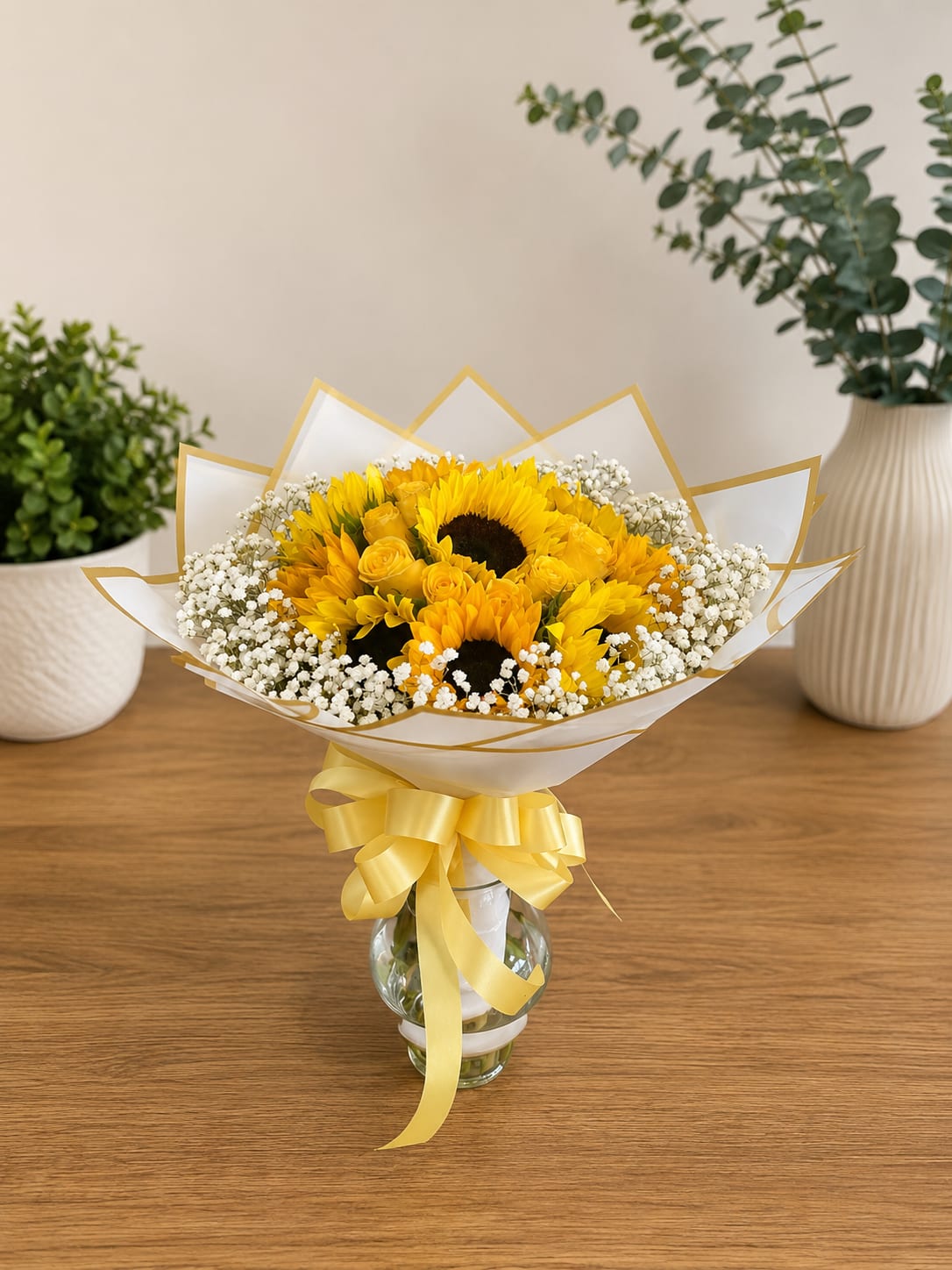     Sun-Kissed Elegance - Bright yellow sunflowers take center stage in this cheerful bouquet, a perfect birthday bouquet or everyday gift to brighten someone's day. Sun-Kissed Elegance is crafted to make the recipient feel warm and celebrated: clustered sunflowers paired with small yellow roses and airy white filler flowers create a sunny, uplifting display ideal for birthdays, congratulations, or just-because moments.  Arranged to feel light and tidy, the stems arrive in a clear glass vase and are finished with white paper edged in gold and a large yellow ribbon for a polished presentation. The mix of yellow, white, and green offers strong visual contrast and soft texture-bold sunflower faces, romantic rose buds, and delicate white accents-making it a ready-to-display centerpiece about the size of a standard vase arrangement. For best enjoyment, keep the vase filled with fresh water and trim stems occasionally to prolong bloom life.