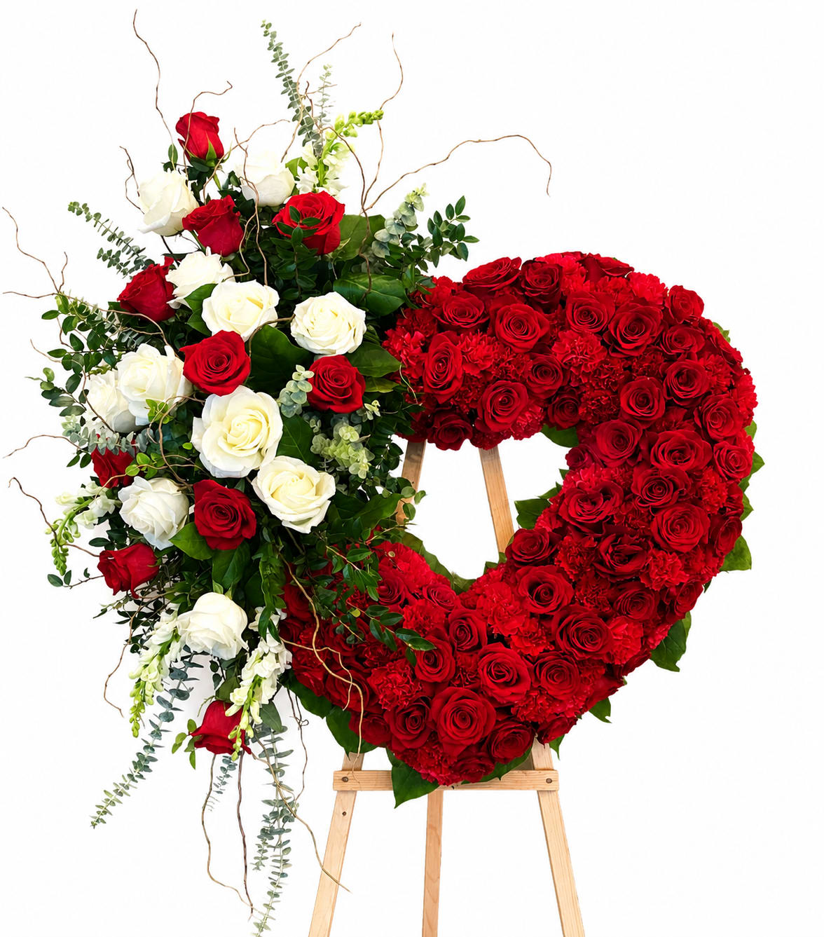 Love Never Fades - A lush heart of deeply red roses creates the first impression-bold, romantic, and unmistakably heartfelt-making this funeral flower arrangement and sympathy tribute a striking way to honor a loved one or to express enduring love. The design reads immediately as a heart-shaped spray for a memorial or a love-themed tribute, so the person you're thinking of will feel deeply remembered and cherished at a service or graveside.  This arrangement pairs the dense 18" heart of red roses with a complementary spray of red and white roses and carnations, set against ribbons of trailing greenery and curving branches that add height and graceful movement. The contrast of crimson blooms with crisp white flowers and fresh green foliage gives the composition formal presence and textured depth. Mounted on a sturdy wooden easel, it arrives ready for display.  Appropriate for sympathy, memorial services, or any occasion where a timeless declaration of love is needed, this piece conveys respect and lasting affection. The heart measures approximately 18" across and is arranged for immediate presentation; light misting of the stems and placement out of direct sun will help extend freshness during services.