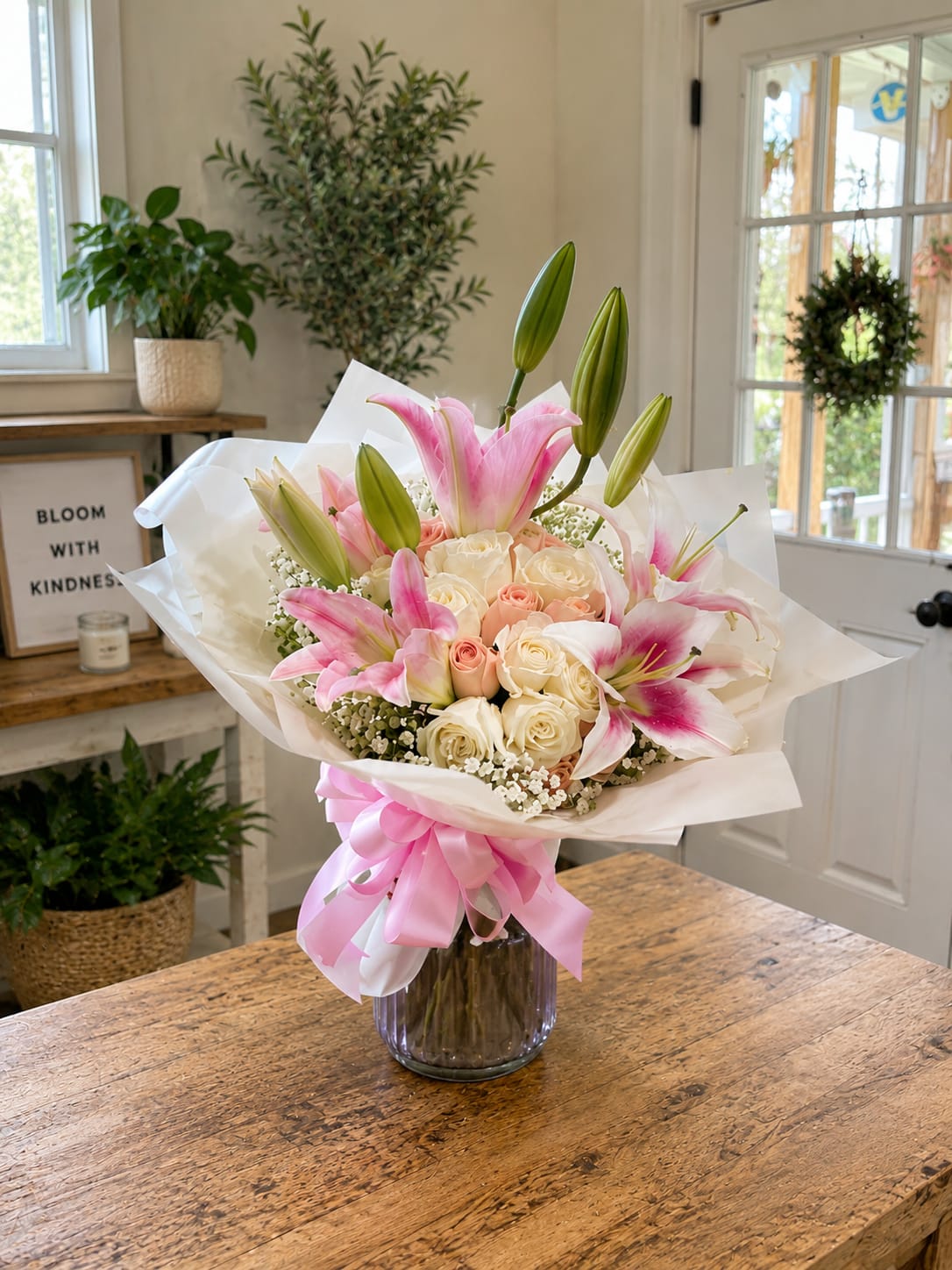 Blushing Elegance Bouquet - Soft pink stargazer-style lilies and cream-to-blush roses create an immediate, romantic impression-perfect as a birthday bouquet, anniversary roses, or a thoughtful thank-you arrangement. The mix of pink, white, peach, and fresh green foliage reads like spring in bloom, so the person you're thinking of will feel celebrated and gently uplifted the moment they see it.  Arranged in a clear glass vase, the flowers are wrapped in white paper and tied with a wide pink ribbon for a polished, gift-ready presentation. Texturally rich lilies and velvety roses give contrast and a light, sweet fragrance; the color palette (pink, white, green, peach) makes this a versatile everyday floral gift. Sized to sit on a table or mantel, it arrives ready to display-place in fresh water and trim stems if desired to keep blooms looking their best for days.