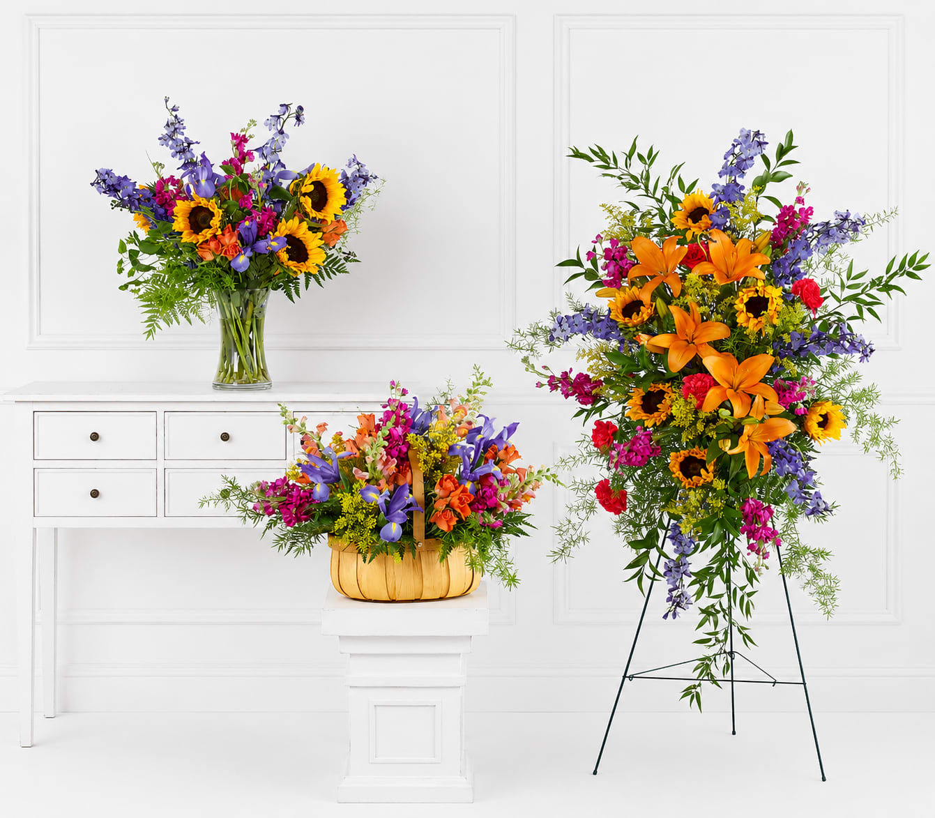 Golden Horizon Bundle - Bright, bold sunflowers and orange lilies create an uplifting focal point in this flower arrangement bundle-perfect as a sympathy or funeral tribute that still celebrates a life with warmth and color. The Golden Horizon Bundle pairs those sunny blooms with purple snapdragon spikes, pink alstroemeria accents, and airy greenery to deliver a vibrant yet respectful display that will make the recipient feel genuinely remembered and honored.  Arranged three ways for flexibility, the bundle includes a clear glass vase arrangement for tabletops, a woven basket design for a homey presentation, and a taller standing piece on a metal easel for services or memorials. Textures range from the sunflower's broad, golden faces to the lilies' graceful petals and the snapdragons' vertical rhythm; sizes vary so the group reads as abundant whether placed on a white pedestal, a table, or near an altar.  Choose this set when you want a brighter, comforting expression of sympathy that balances cheer with reverence-appropriate for families seeking a hopeful tone at a funeral or memorial. Standard care: keep vase water fresh and trim stems every few days; if delivered for a service, specify placement preferences and we'll coordinate the presentation to fit the venue.