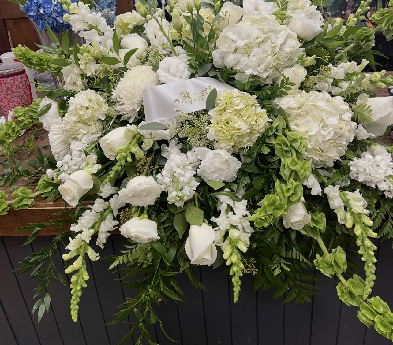Casket cover - A sweeping, low casket cover in serene white and fresh green makes a quiet, polished statement for a funeral service or memorial. This sympathy arrangement combines white hydrangeas, roses, calla lilies, snapdragons, daisies and chrysanthemums for a full, cascading silhouette that reads both formal and comforting; the white satin ribbon across the center adds a personalized, respectful touch.  Densely layered rounded blooms and taller vertical accents create depth and a soft, textured profile that drapes gently over a casket. Trailing greenery and mixed white florals give the piece a natural movement while maintaining an elegant, unified palette. Sized for prominent display, it offers a composed, enduring expression of remembrance and support for the family during a difficult time.