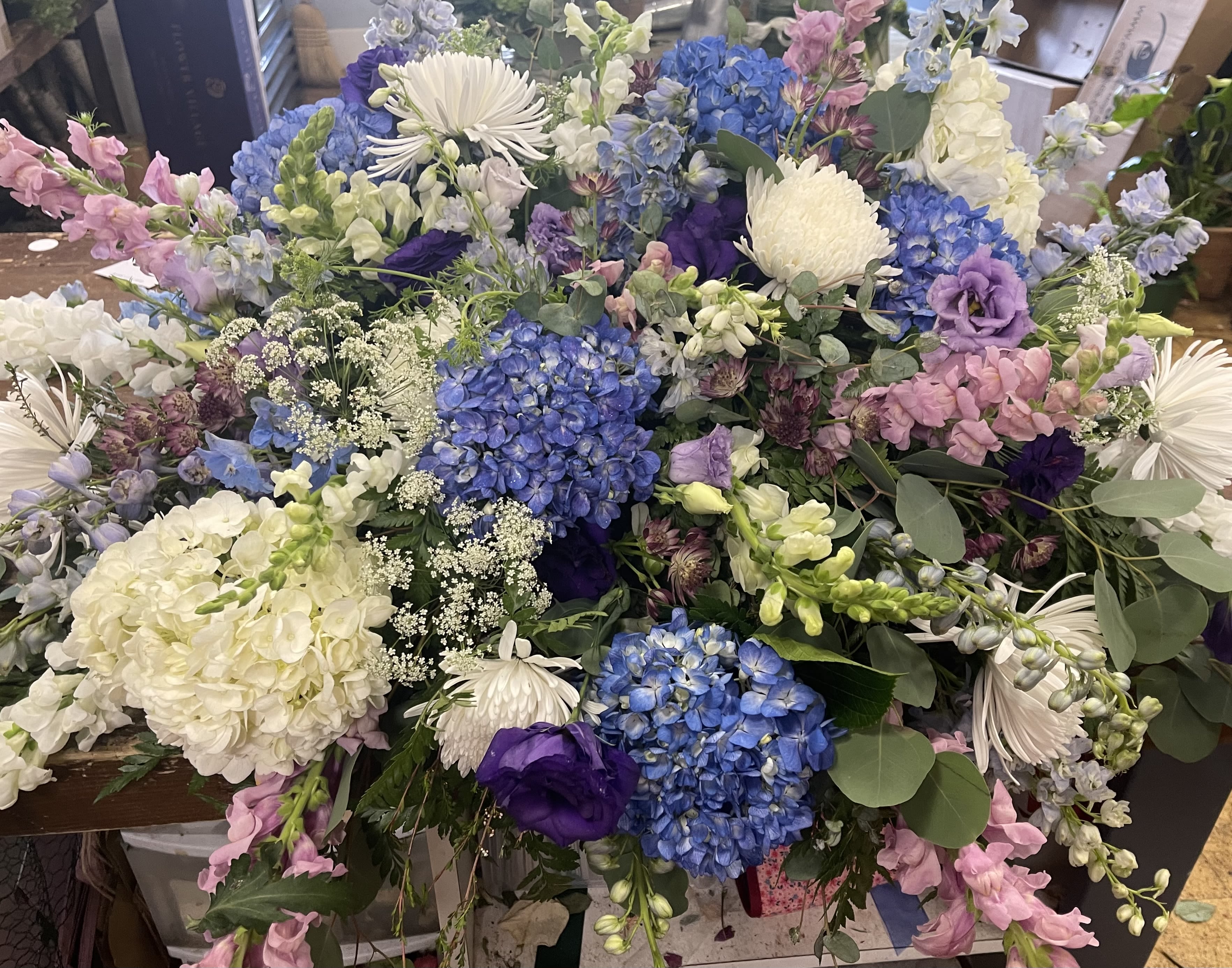 Monet’s Garden - Clusters of powder-blue hydrangeas set the tone in this large, garden-style flower arrangement-perfect as a sympathy or funeral tribute or a gentle Thinking of You gift. The mix of blue, white, pink, and purple blooms creates a soft, comforting palette that reads as both romantic and quietly uplifting, making it appropriate for winter remembrances as well as year‑round moments when you want someone to feel seen.  This abundant bouquet pairs rounded hydrangea heads with airy lisianthus, tall snapdragon spikes, and cheerful white daisies for a lively mix of shapes and textures. Eucalyptus and assorted greenery drift through the design, adding a fresh, silvery contrast and a loose, hand‑gathered silhouette. The overall effect is rustic yet refined-an arrangement that looks freshly cut from a cottage garden.  Whether you're marking a Quinceañera with gentle pastels, honoring Día de las Flores Amarillas with thoughtful contrast, or sending condolences, Monet's Garden conveys warmth and care. Sized to make an impact, it arrives ready to display; simple care-fresh water and a cool spot-will help the blooms last and continue to comfort the person you're thinking of.